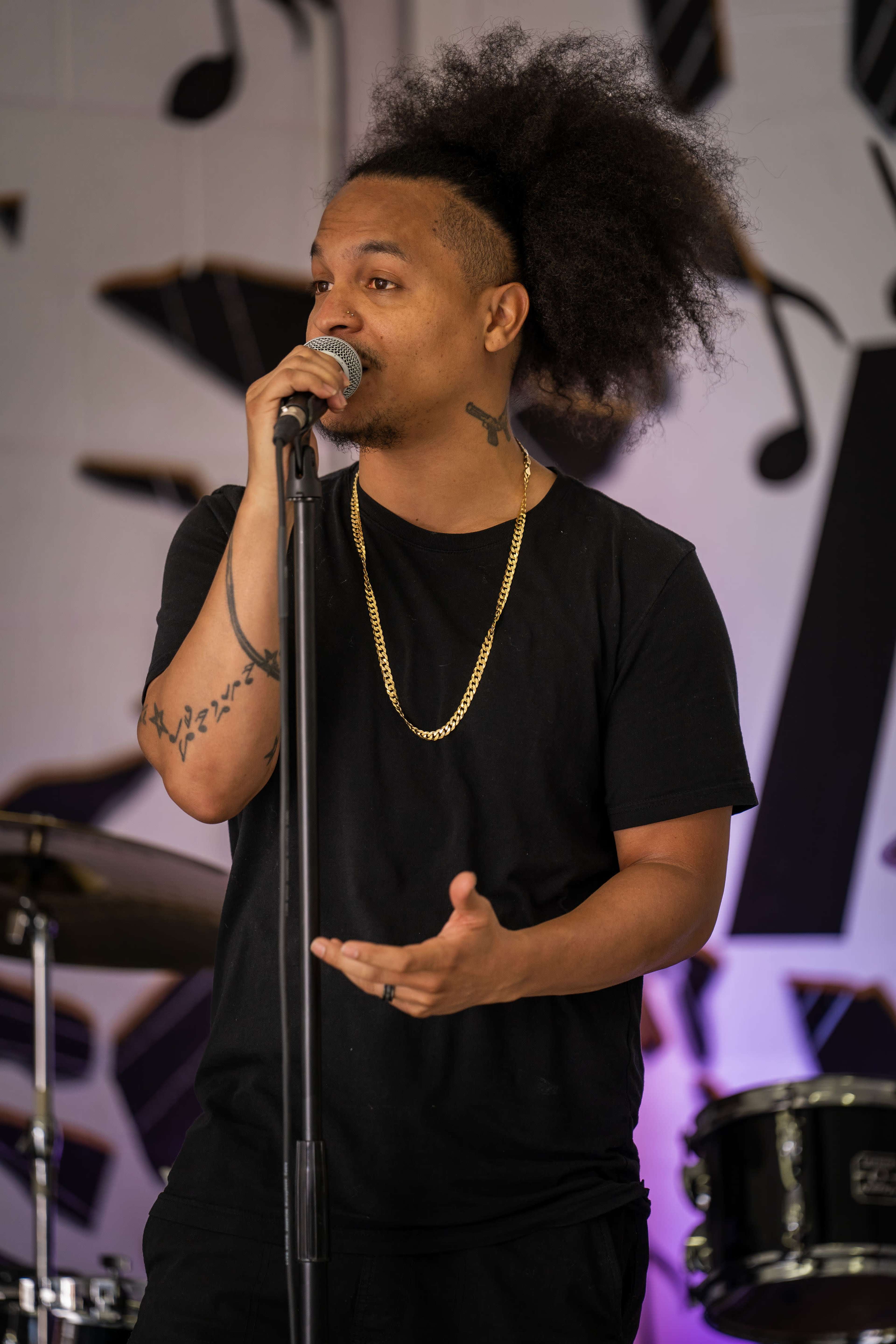A man with a large, curly hairstyle stands at a microphone, speaking while gesturing, in front of a backdrop featuring musical notes.