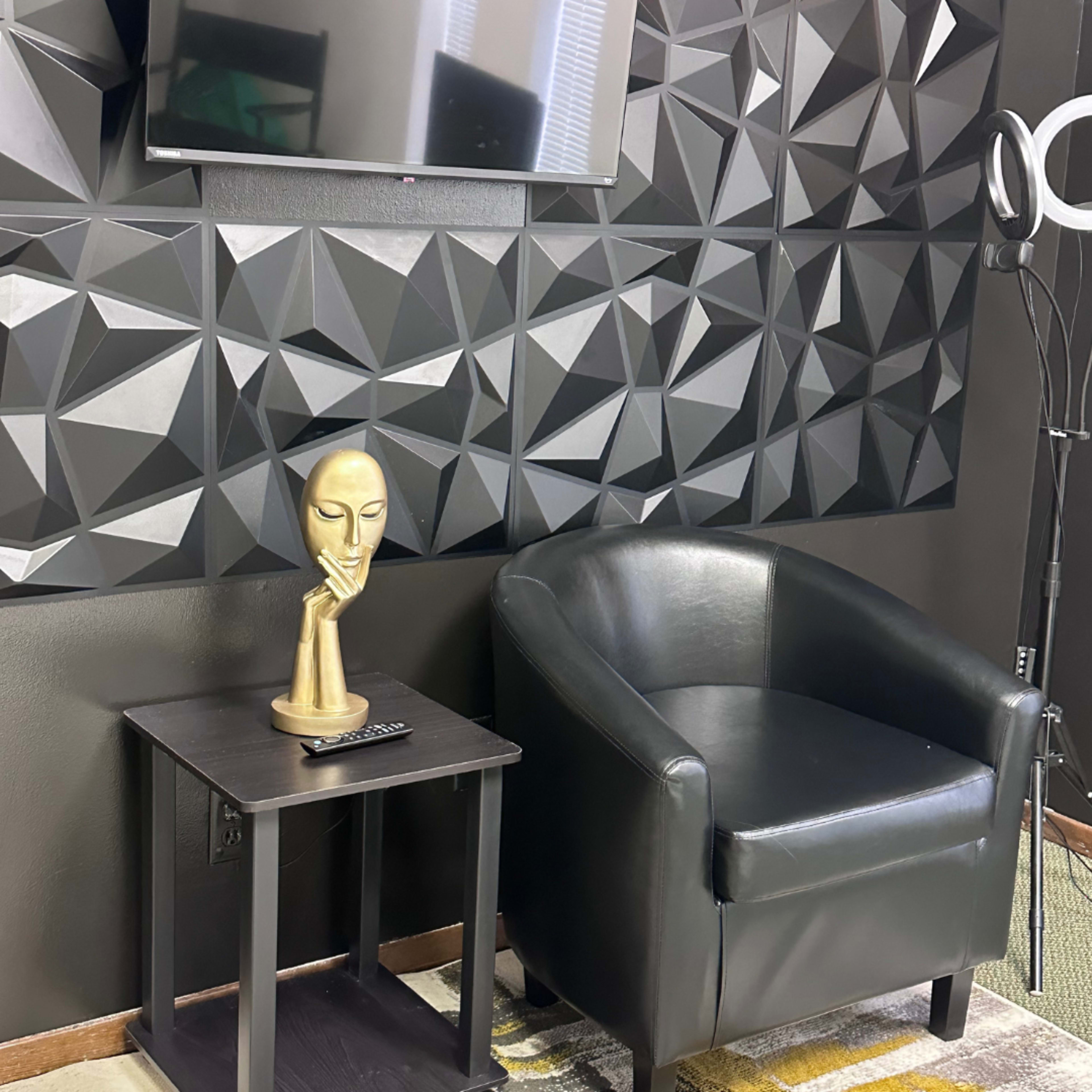 A black chair next to a small table holding a gold sculpture of a head, with a textured black wall and a television above.