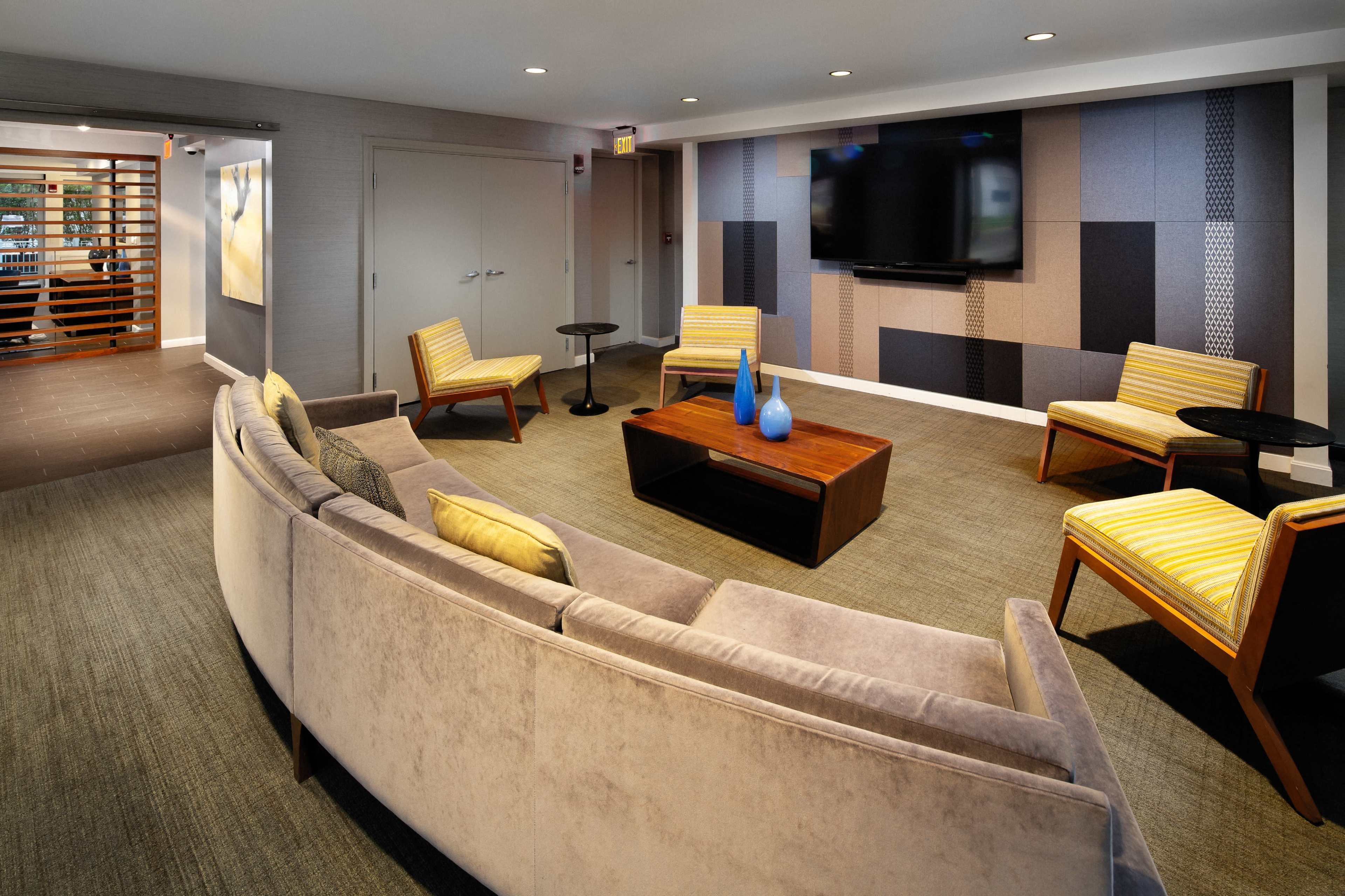 The image shows a modern lounge area featuring a curved gray sofa, several yellow-striped chairs, a wooden coffee table, and a wall-mounted television surrounded by patterned panels.