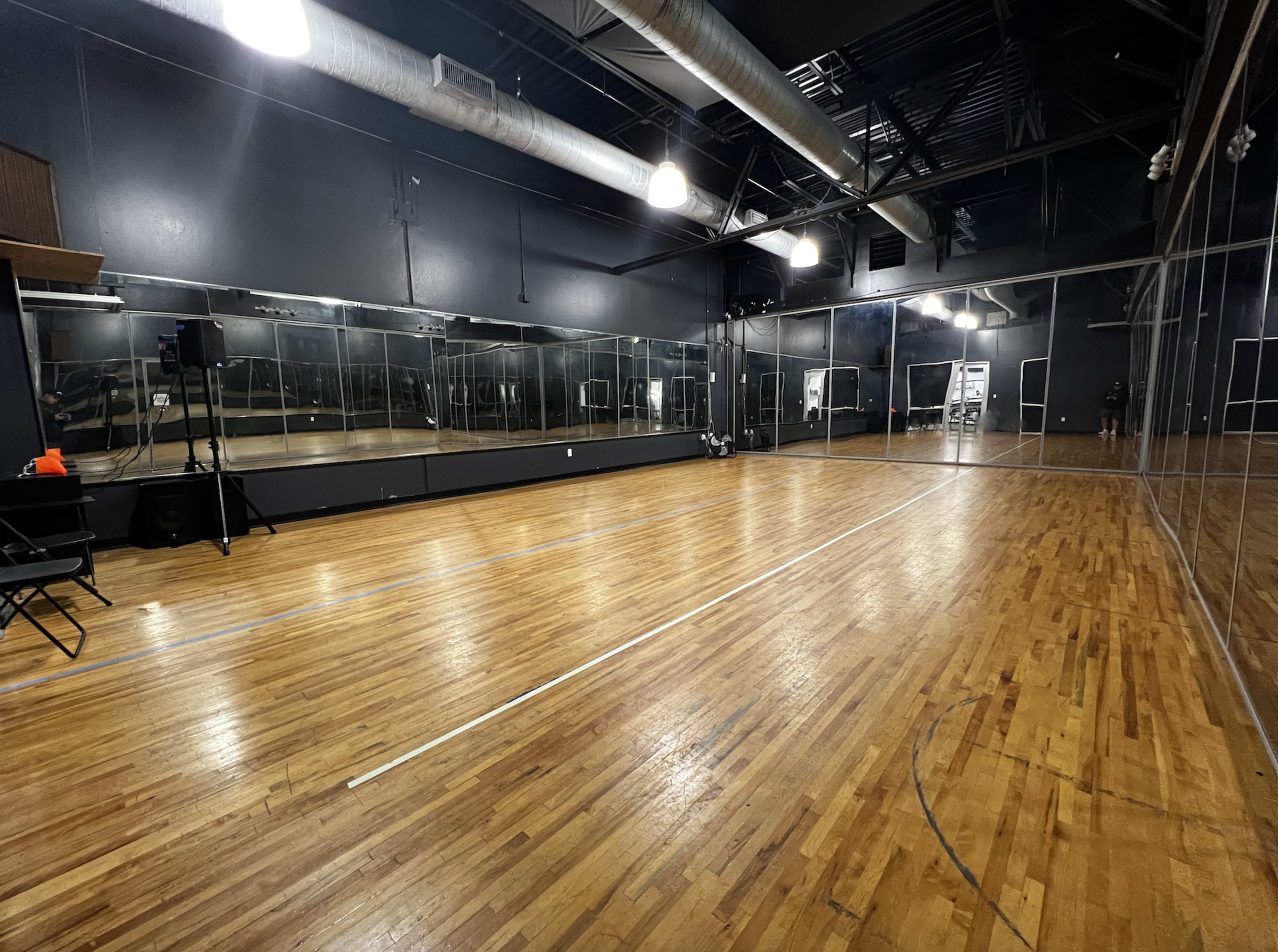 A spacious dance studio with wood flooring, large mirrors along one wall, and overhead lighting.