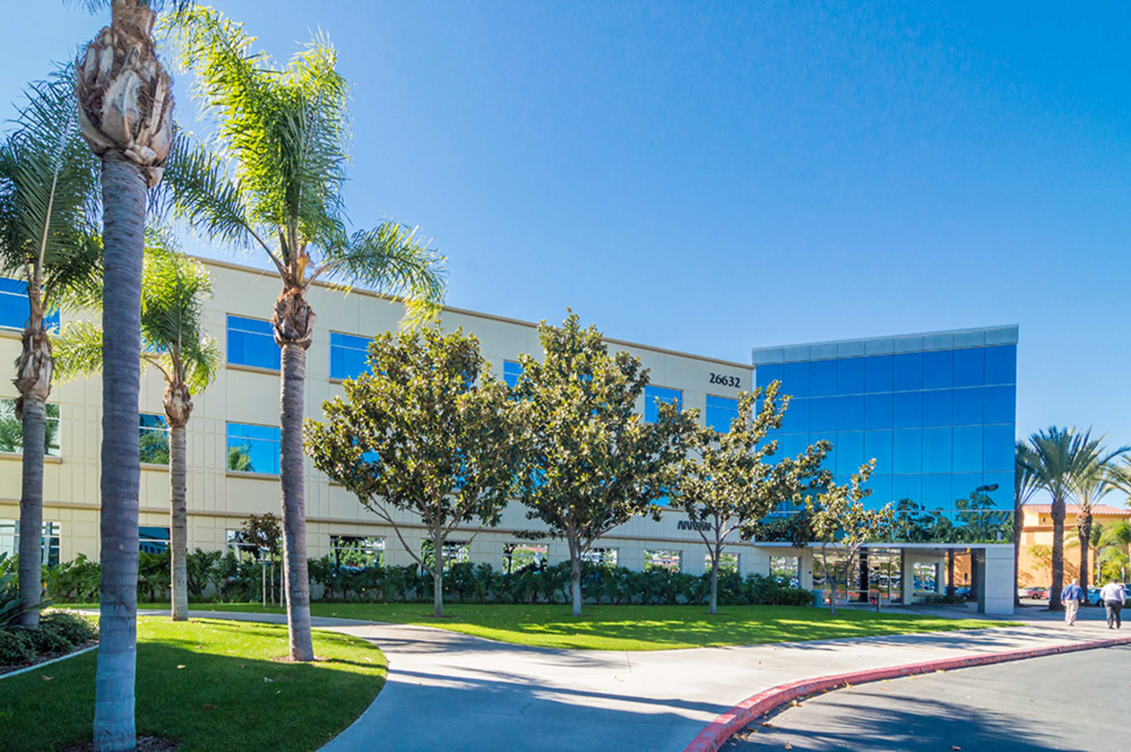 The image shows a modern office building with large glass windows, surrounded by palm trees and landscaped greenery.