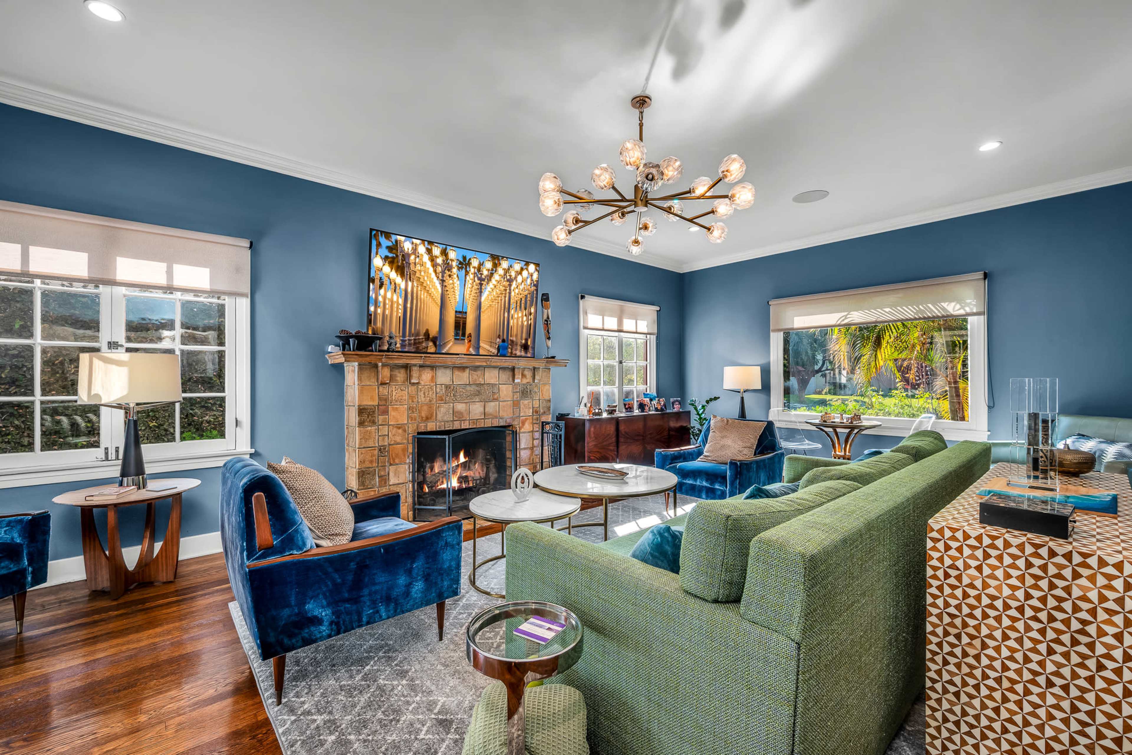 A modern living room features a stone fireplace, a large television, and furniture in blue and green hues, with windows showing a view of greenery outside.