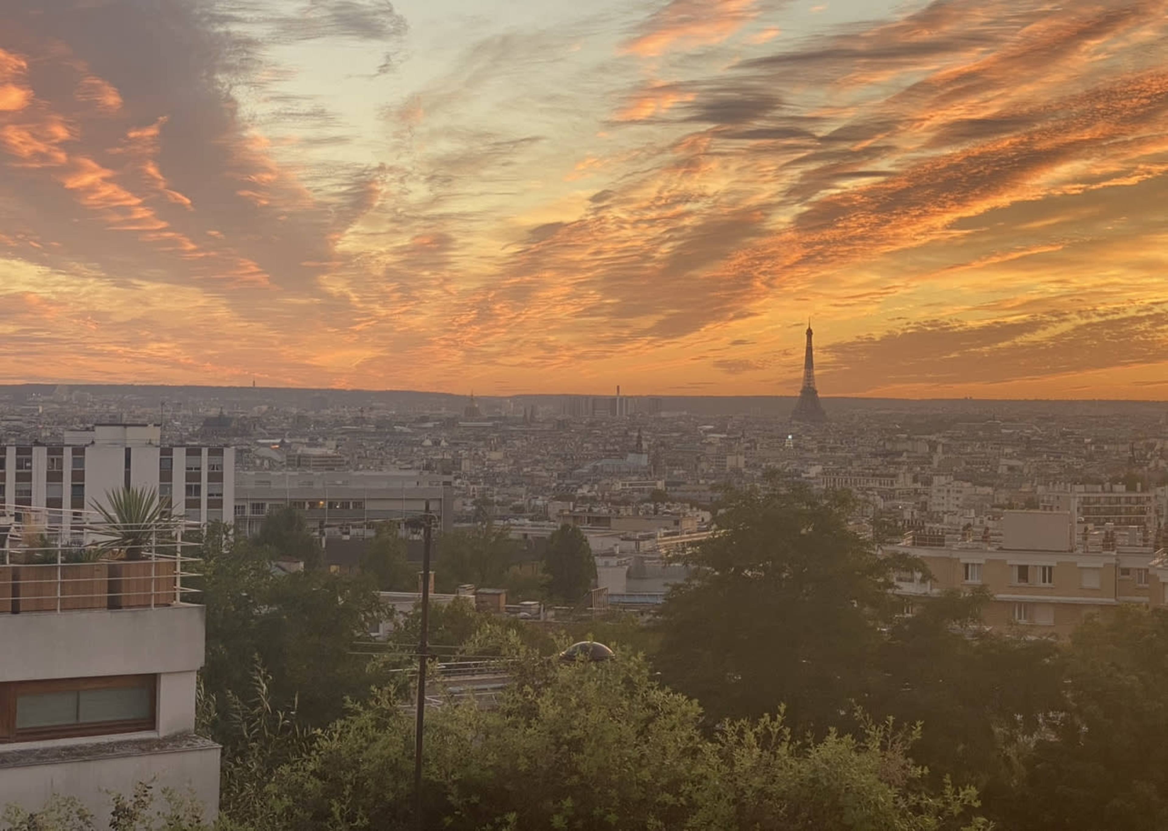 A panoramic view of Paris at sunset, with vibrant clouds and the Eiffel Tower visible in the distance.