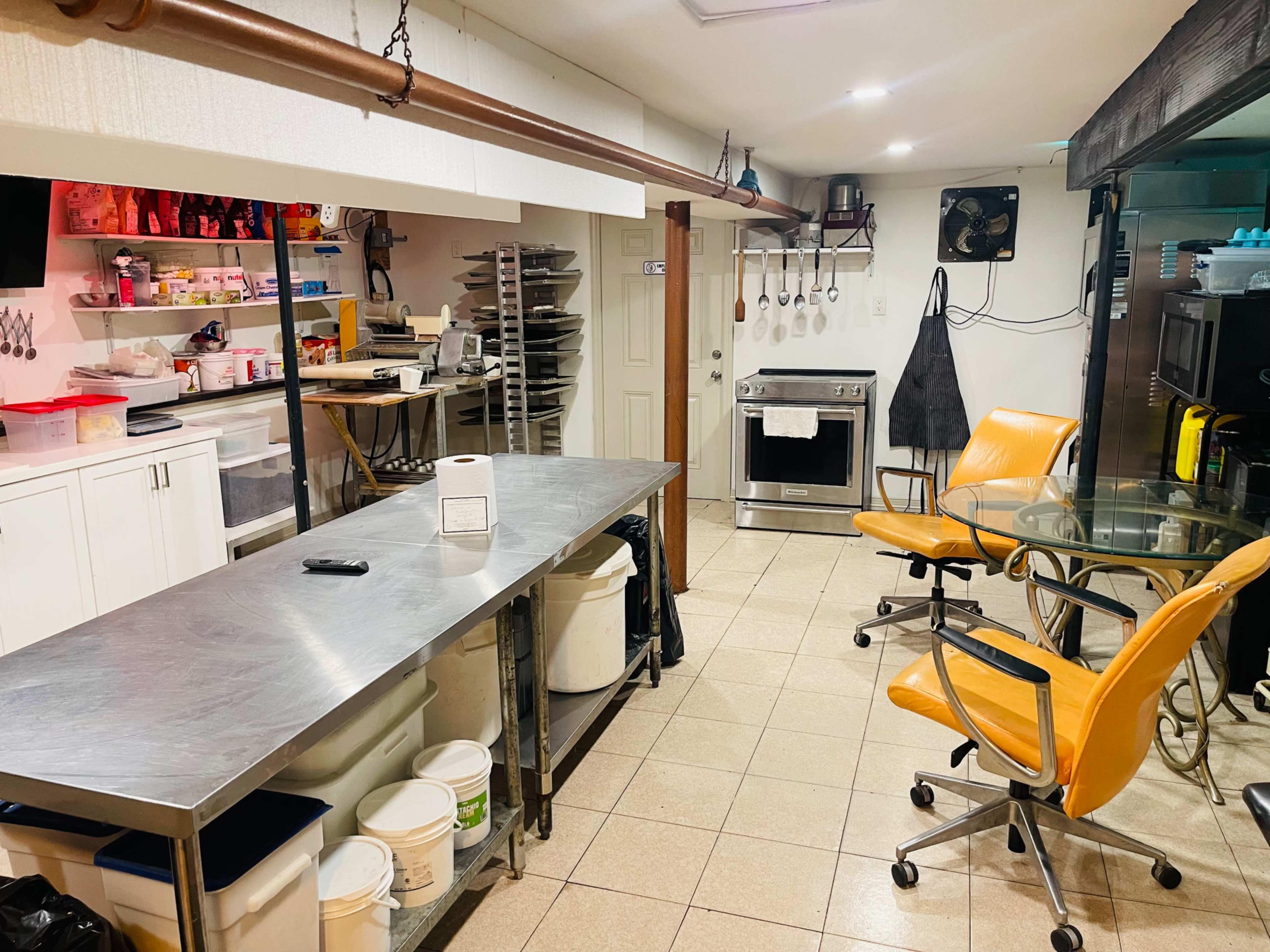 The image shows a well-organized kitchen featuring stainless steel countertops, various kitchen equipment, and an orange chair near a glass table.
