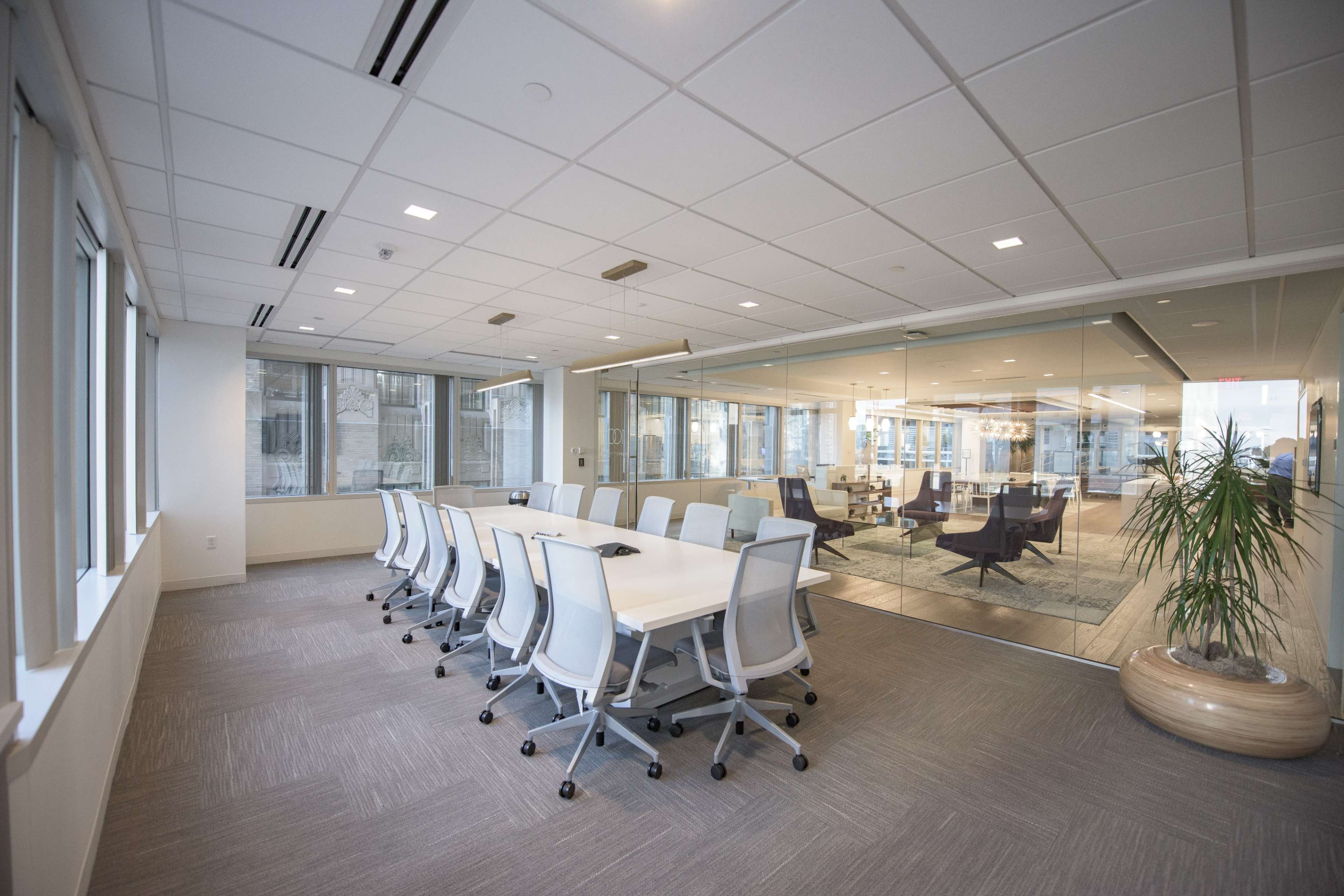 A modern conference room features a long white table surrounded by rolling chairs, with large windows offering natural light and a glass wall separating it from a lounge area.