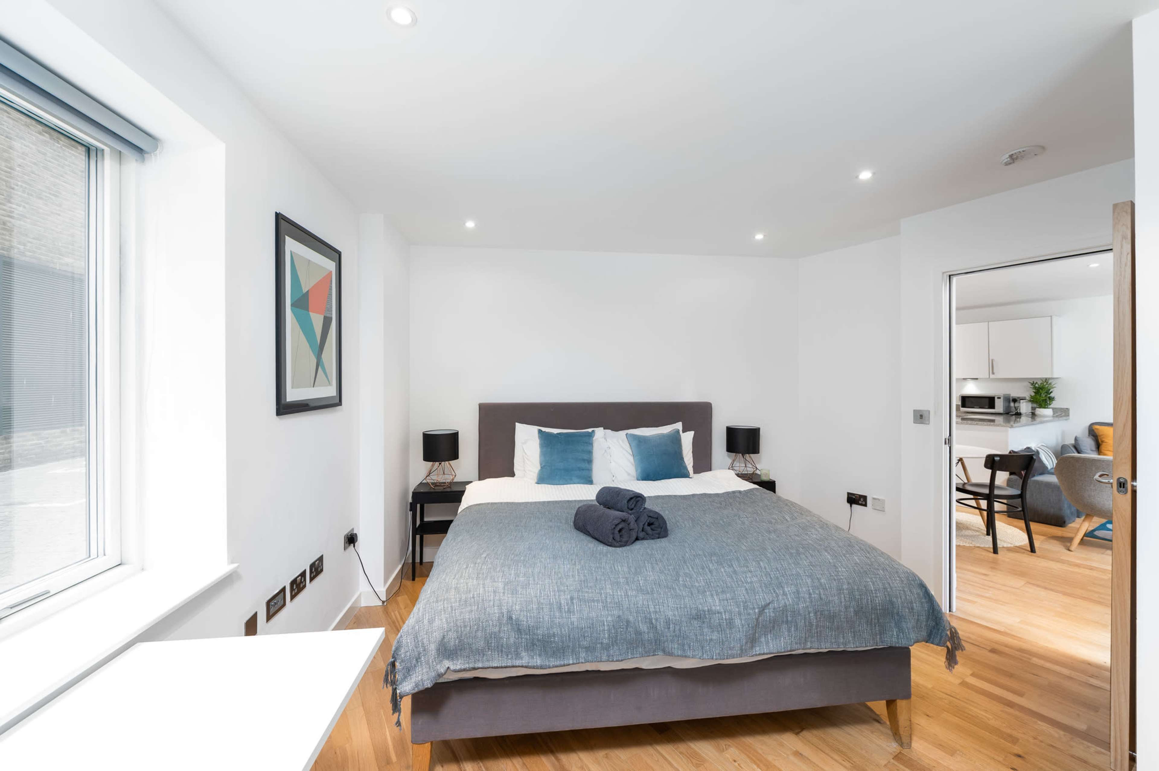 A modern bedroom features a large bed with gray bedding, two bedside tables with lamps, and a window allowing natural light.
