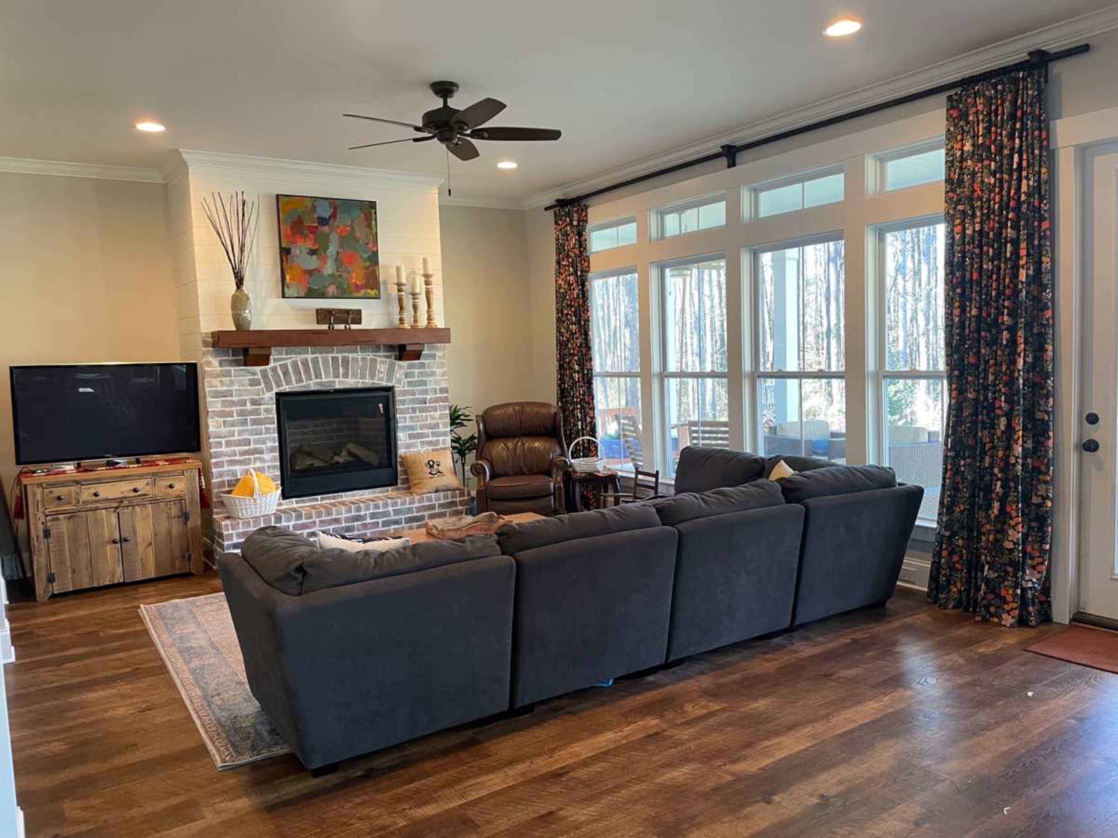 The image shows a cozy living room featuring a sectional sofa, a brick fireplace with a mantel, a television, and large windows with floral curtains.