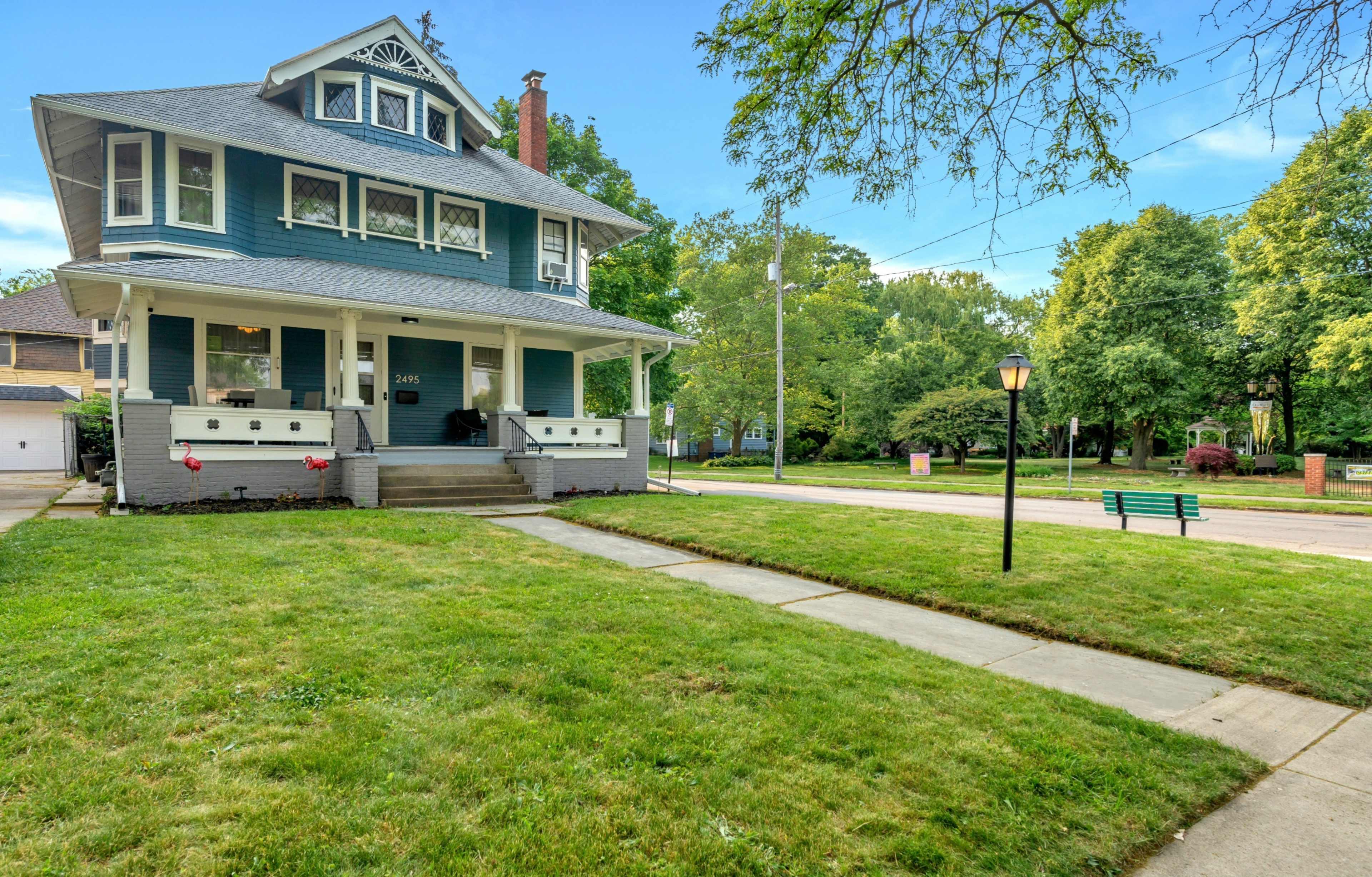 A large, two-story blue house with a front porch and manicured lawn is situated on a tree-lined street.