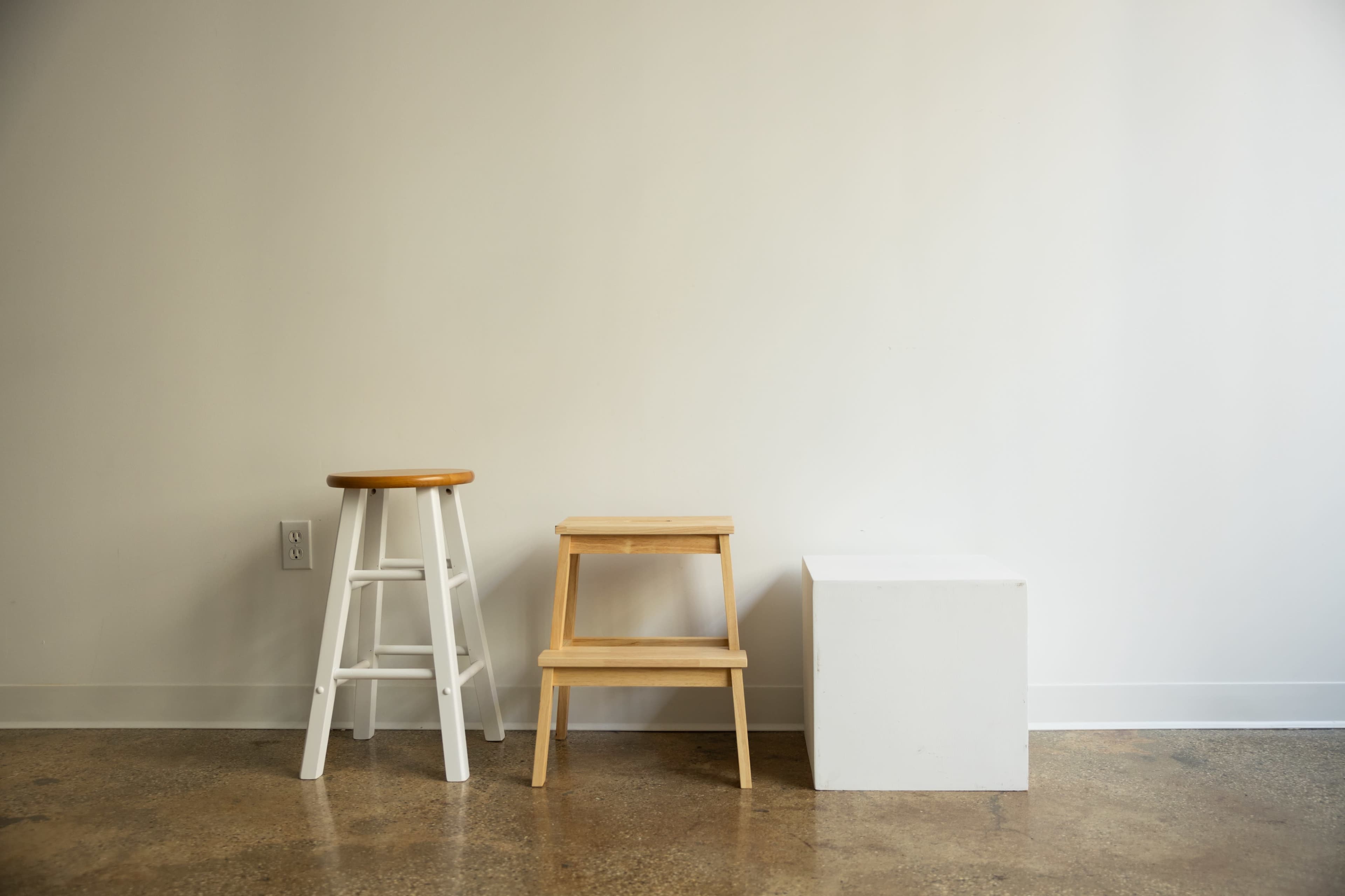 The image shows three different types of stools or stands in a minimalist setting against a plain white wall.