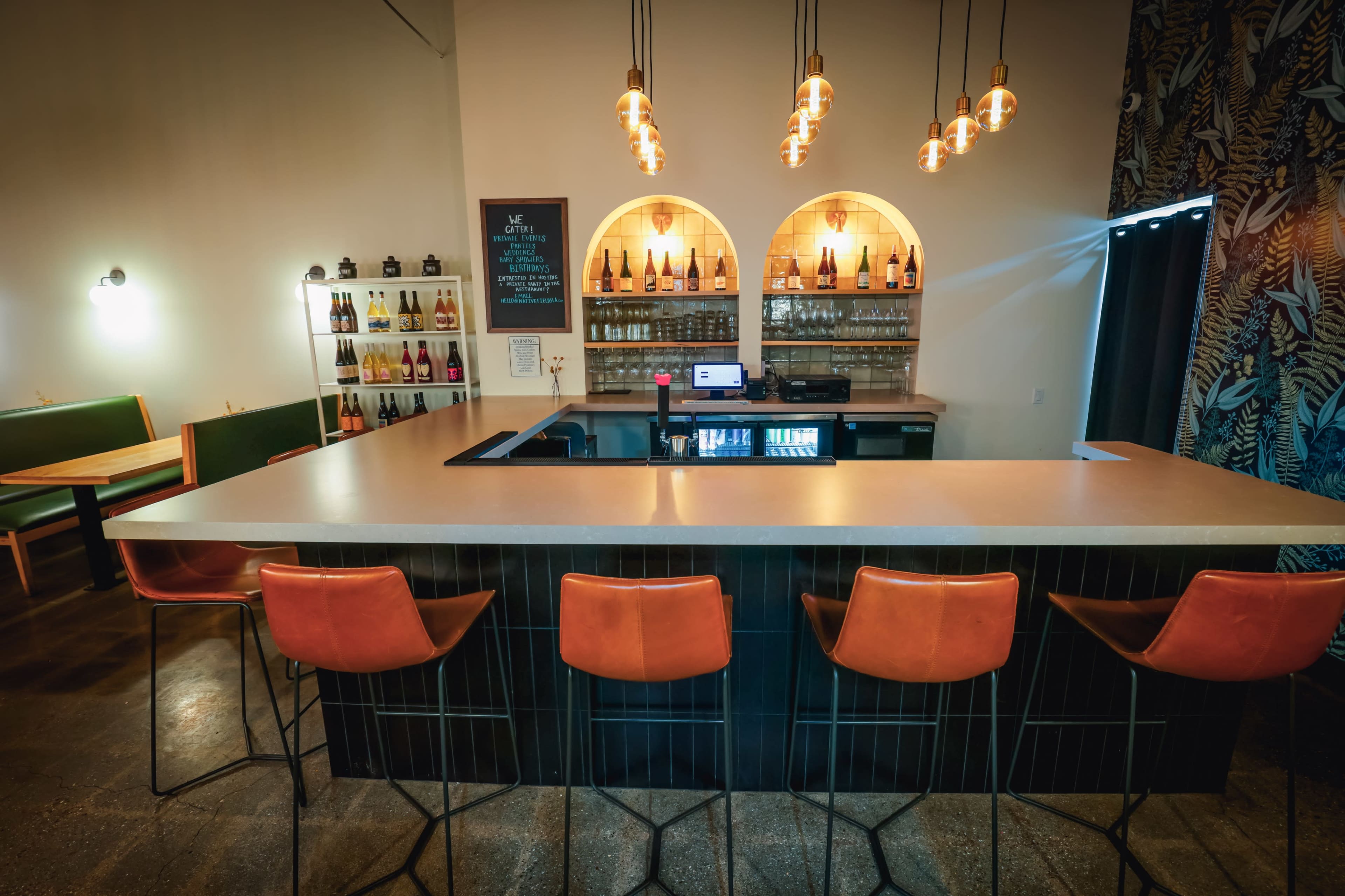 The image shows a modern bar area with four orange bar stools, a countertop, shelves of wine and spirits in the background, and decorative lighting fixtures hanging above.
