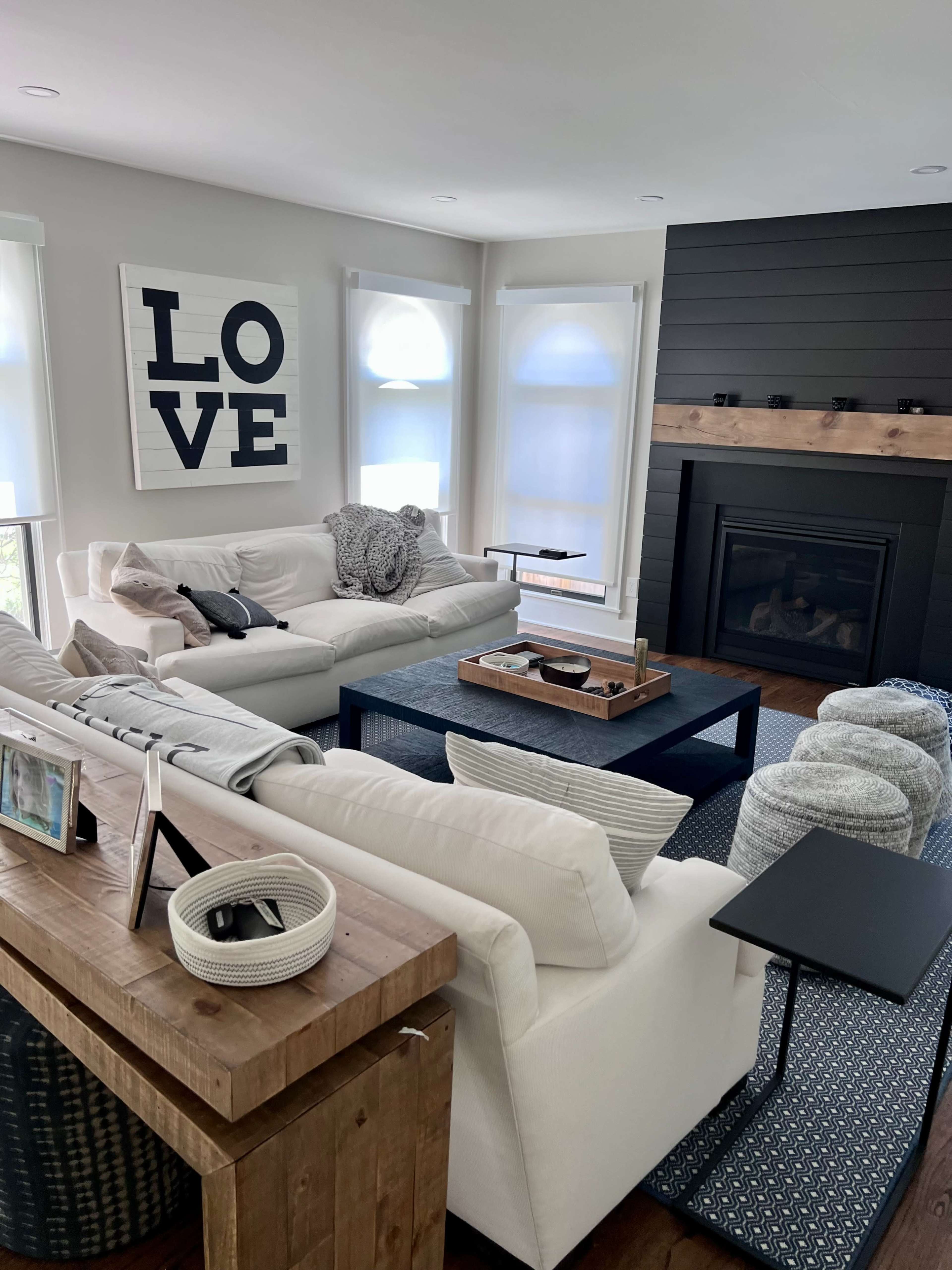 The image shows a modern living room with a white sectional sofa, a dark wood coffee table, and a fireplace surrounded by dark shiplap walls, adorned with a large wall art that says "LOVE."
