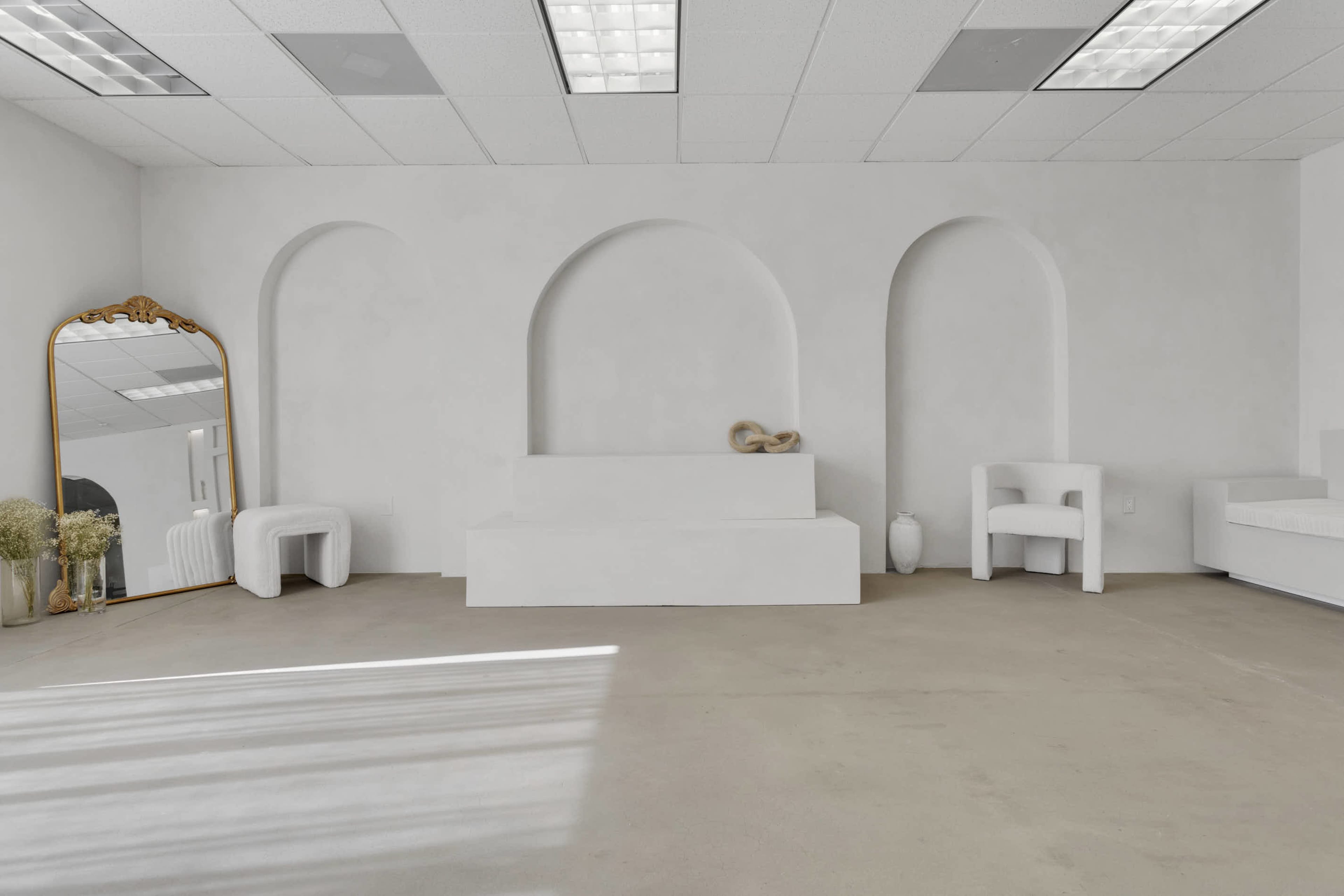 The image shows a minimalist room with arched recesses, a white platform bench, and various decorative objects against a light-colored wall.