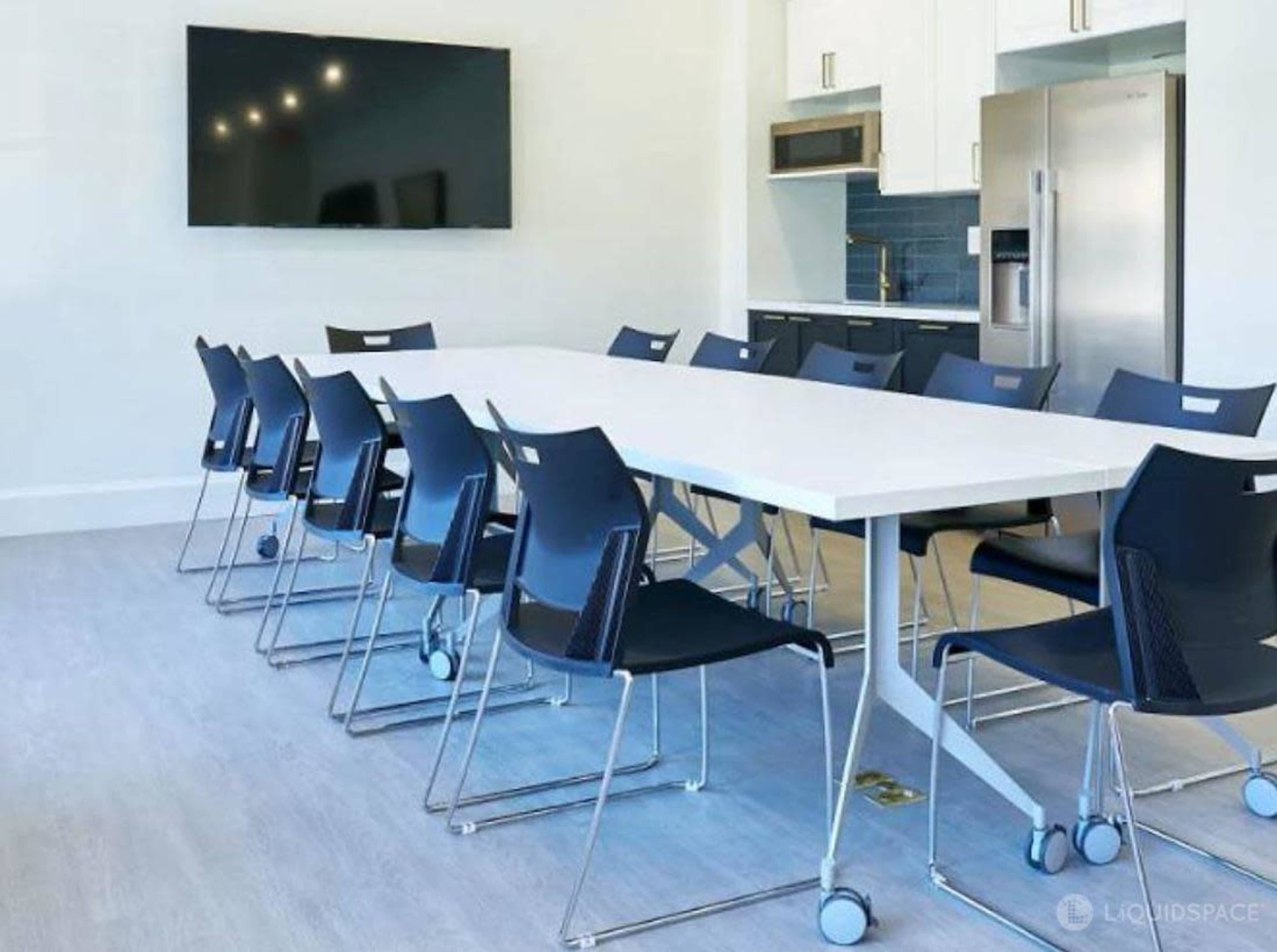 A long white conference table with black rolling chairs is set in a modern meeting room featuring a large screen and kitchen area.