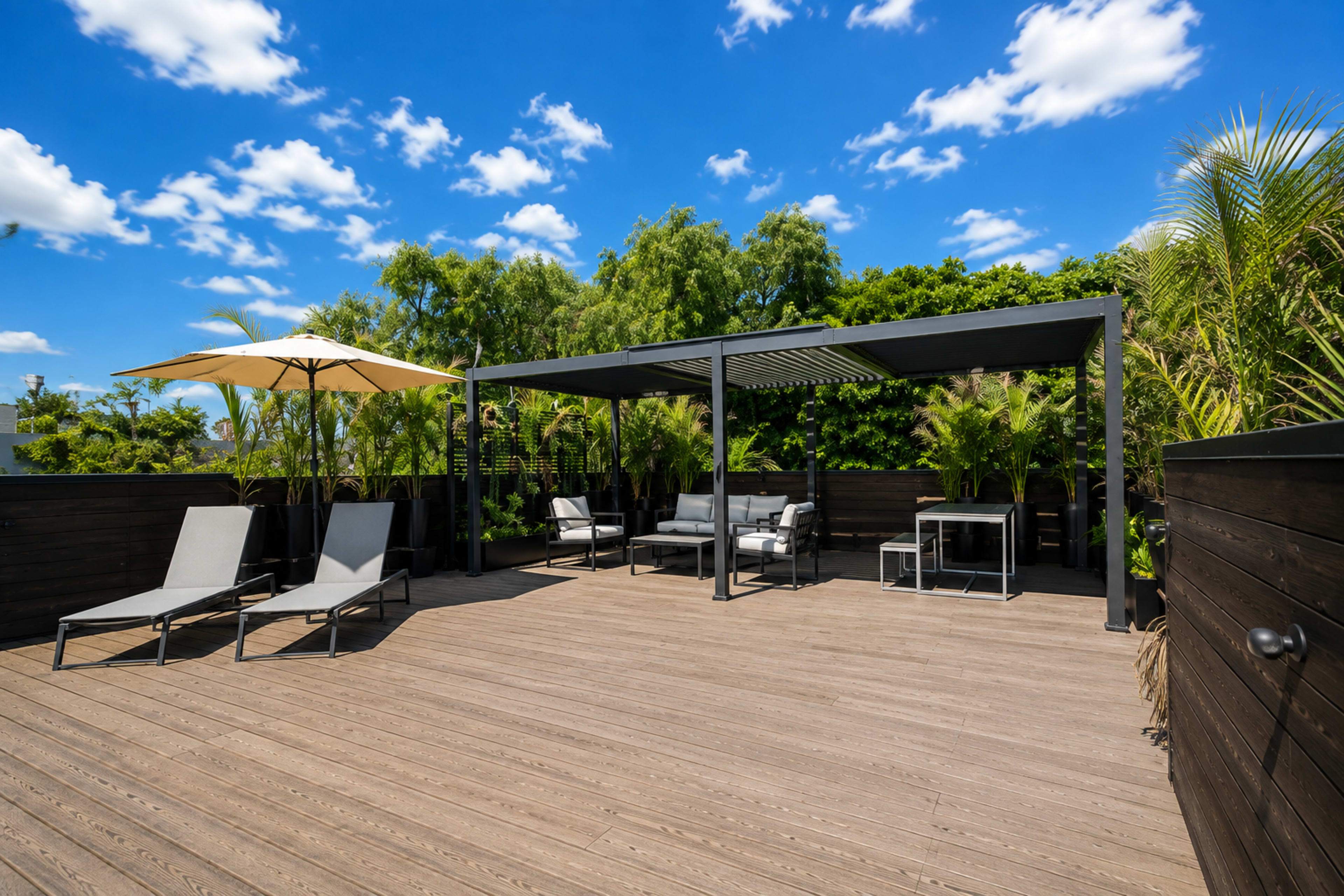 The image depicts a spacious rooftop terrace featuring lounge chairs, a shaded seating area, and tropical greenery under a bright blue sky with scattered clouds.
