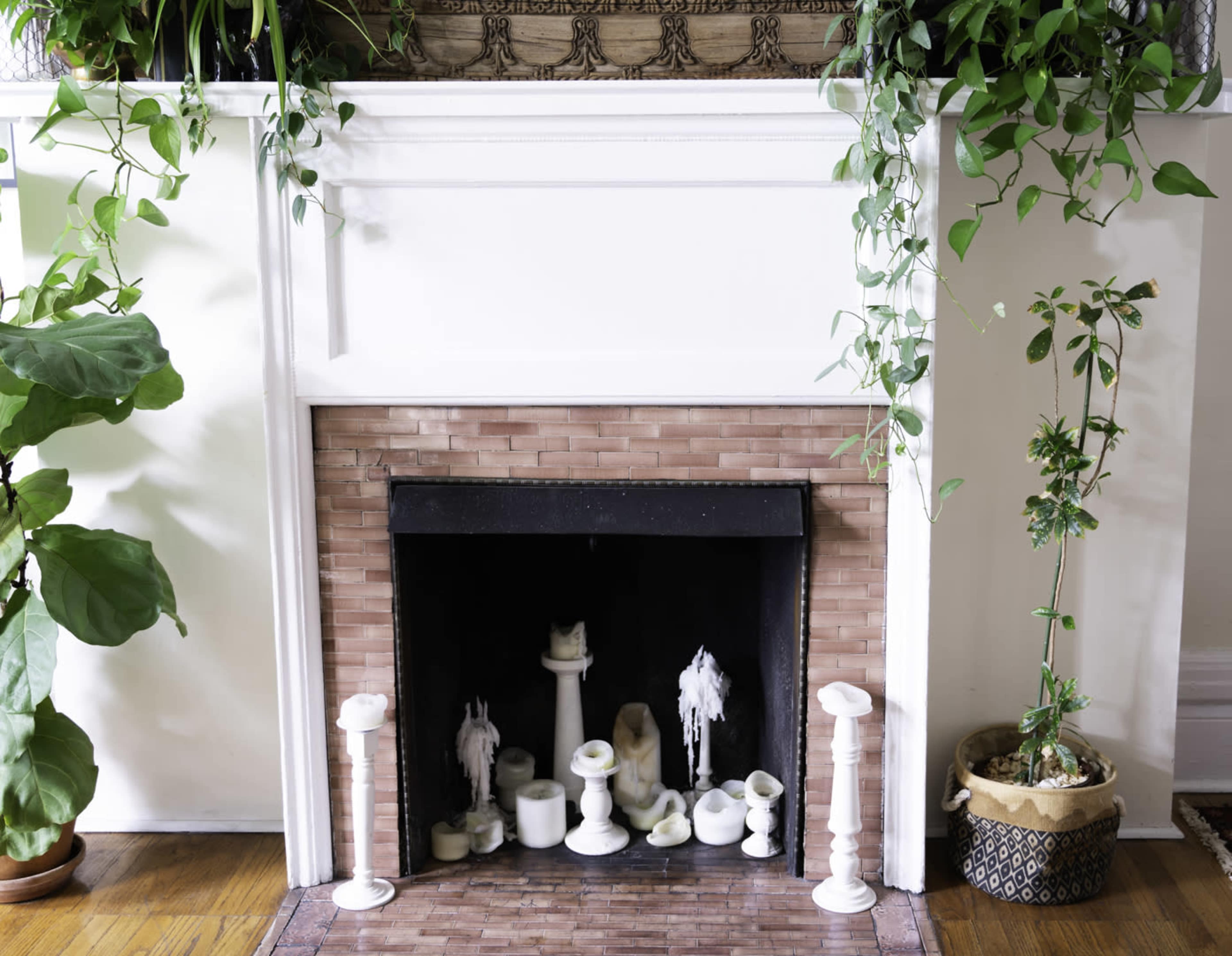 A white fireplace with a textured stone front, surrounded by green plants and decorated with various candles and a small statue.