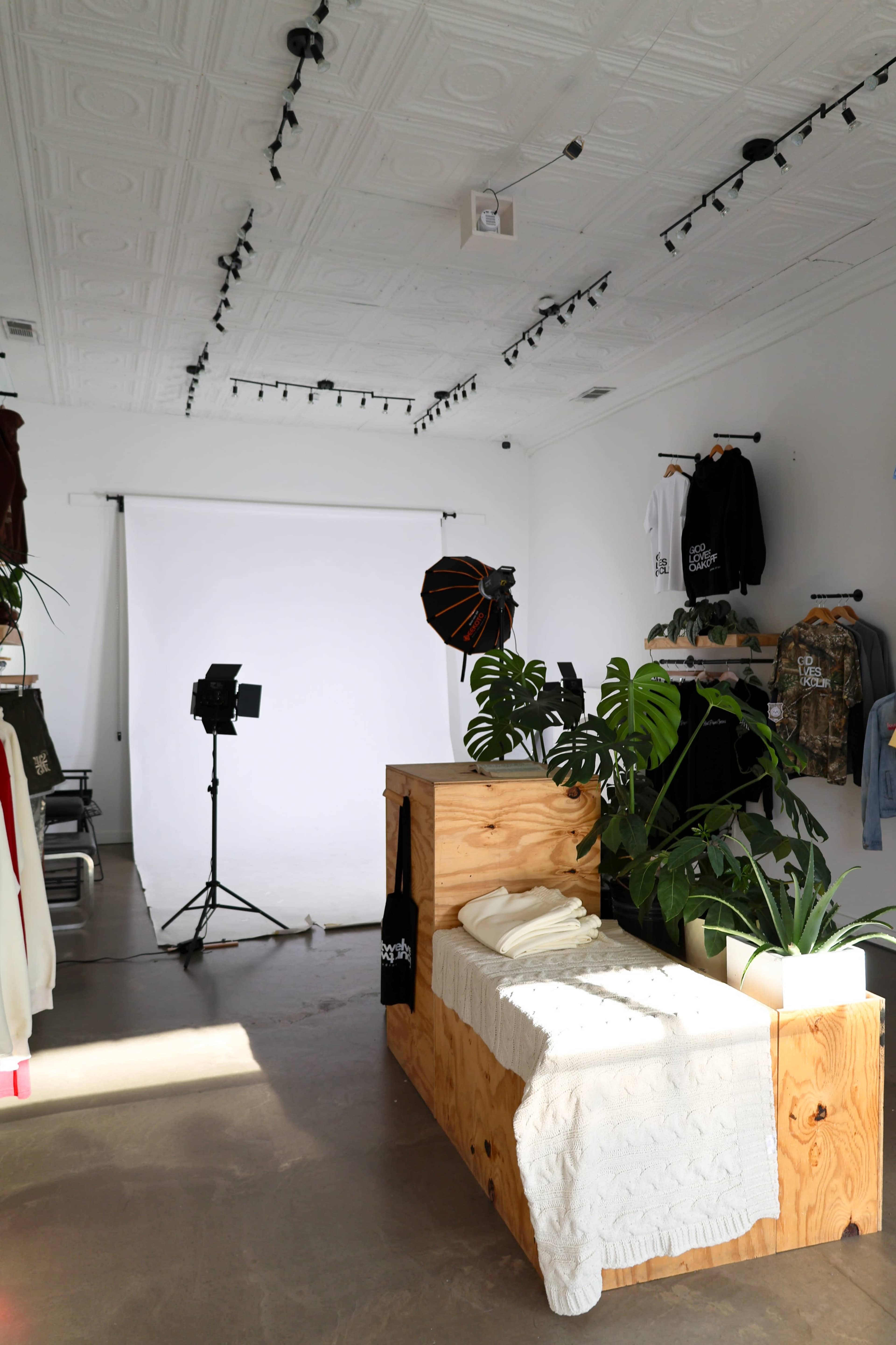 The image shows a minimalist clothing store interior featuring a wooden display with a bed, plants, and a photography setup in front of a white backdrop.