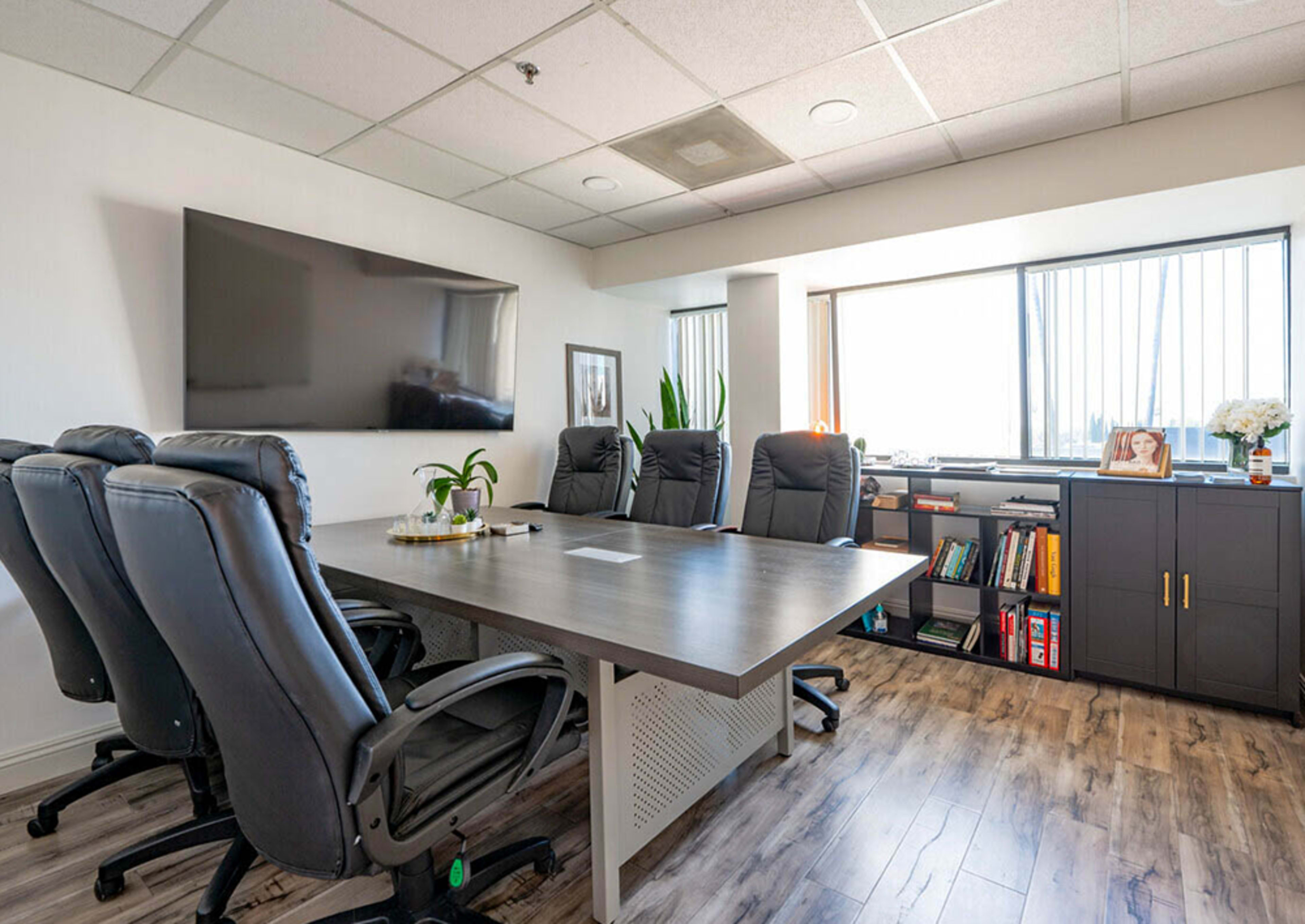 The image shows a modern office meeting room with a large table, black leather chairs, a wall-mounted TV, and a window containing natural light.
