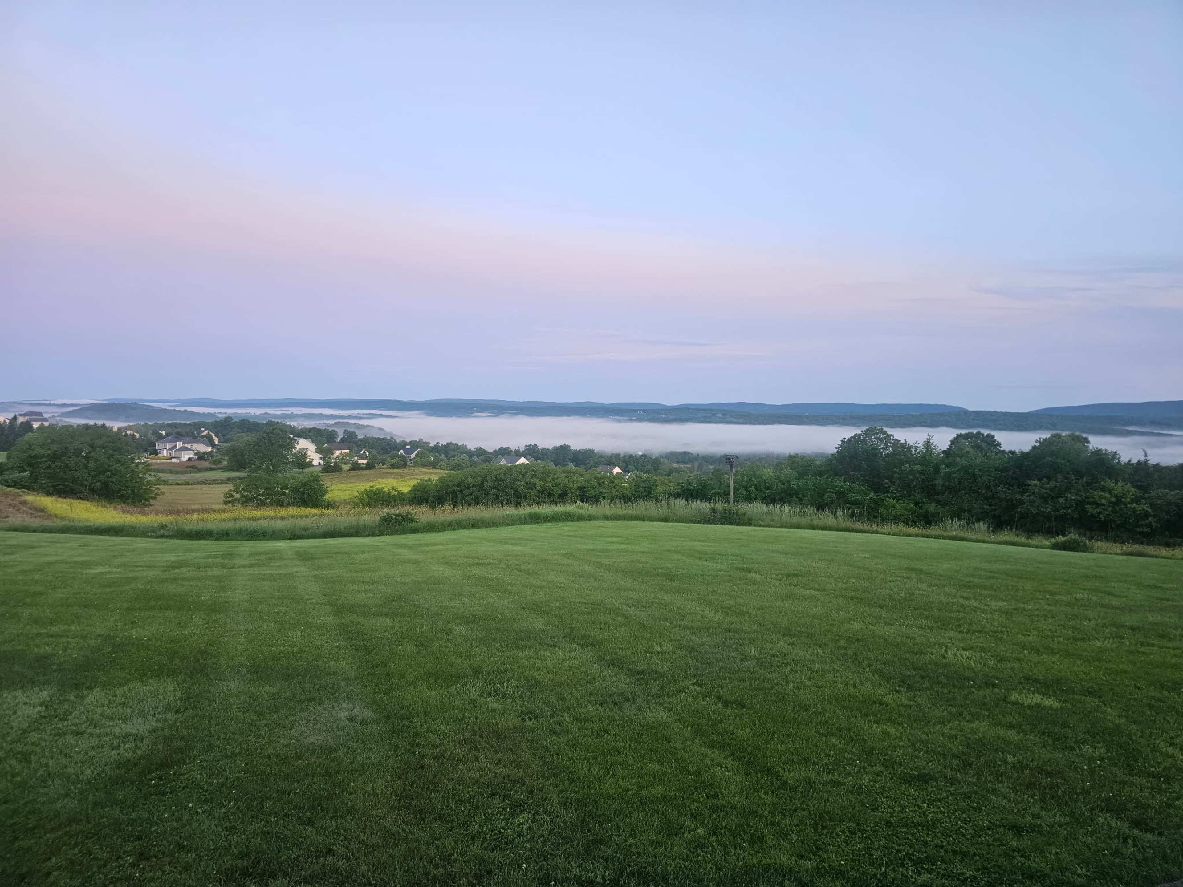 The scene features a grassy landscape overlooking a fog-covered valley and rolling hills under a pastel sky.