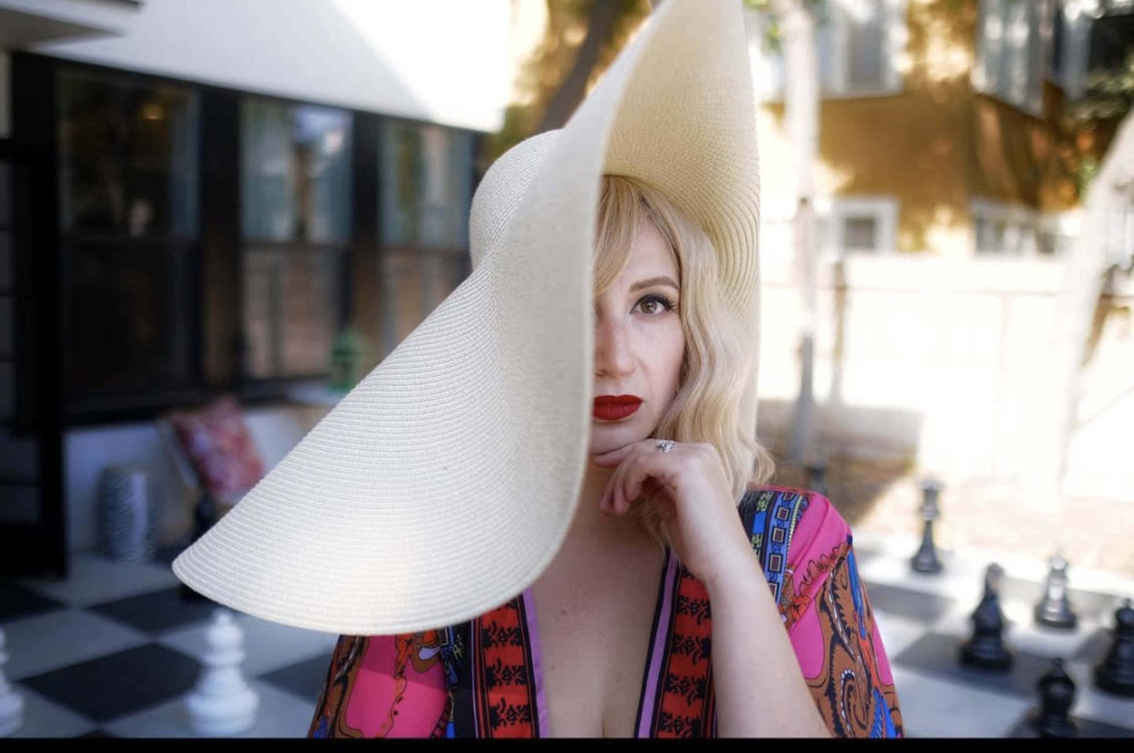 A woman with blonde, wavy hair wears a large straw hat and poses thoughtfully outdoors, with a chessboard in the background.