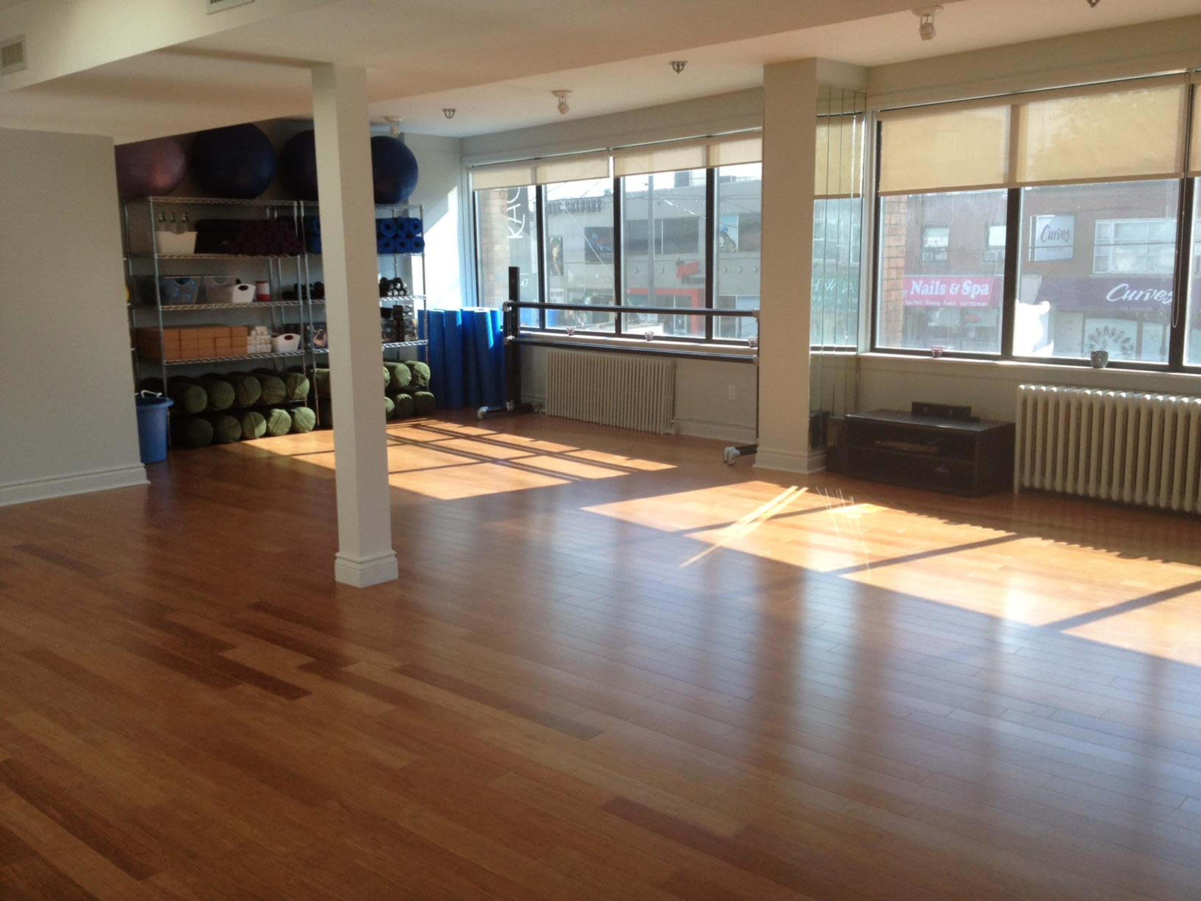 A spacious, well-lit exercise studio with wooden flooring, large windows, and fitness equipment neatly arranged against the walls.