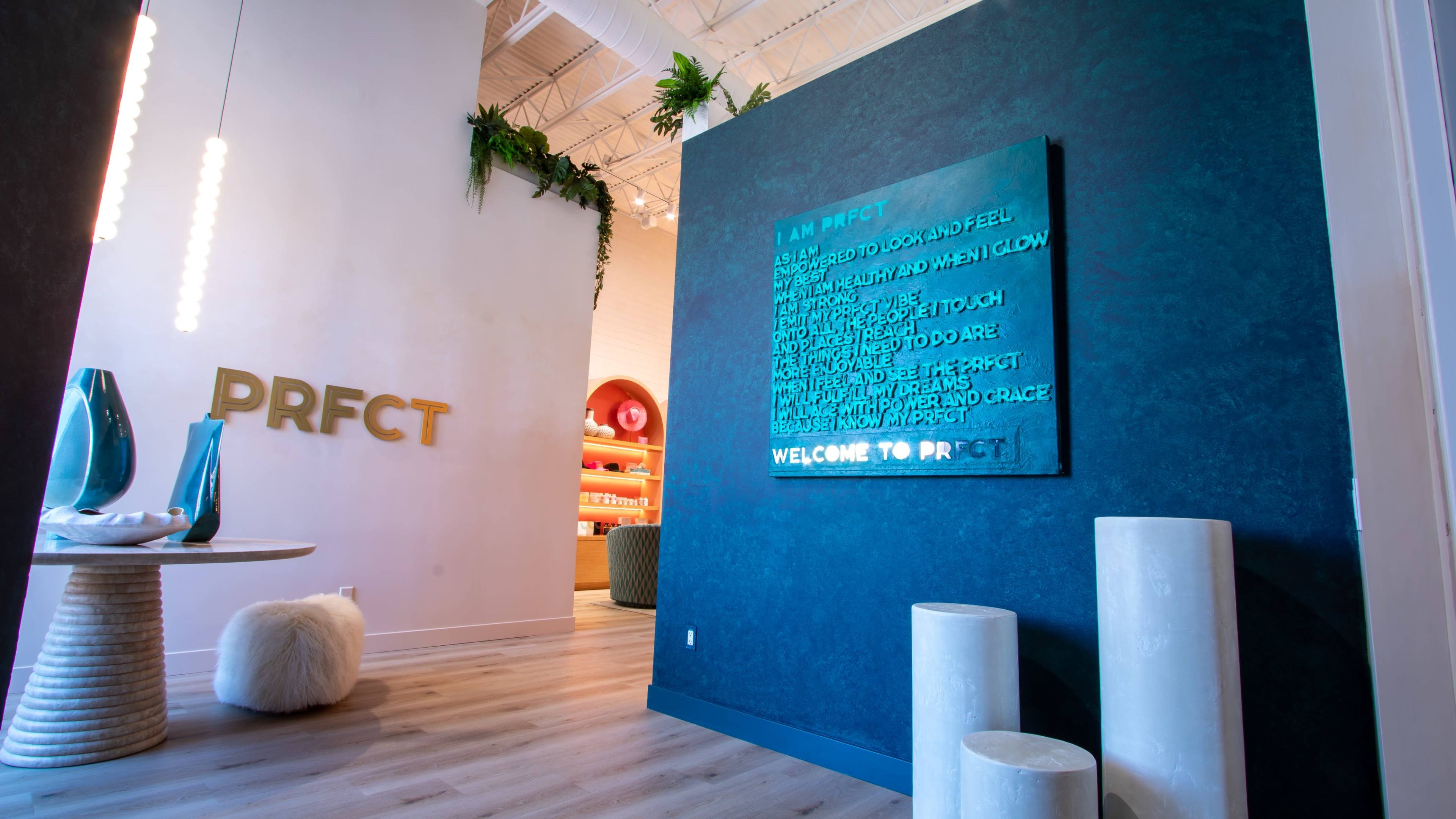 The image shows a modern interior space featuring a wall with the text "I AM PRFCT," a round table with decorative items, and a welcoming entrance with the word "PRFCT" displayed prominently.