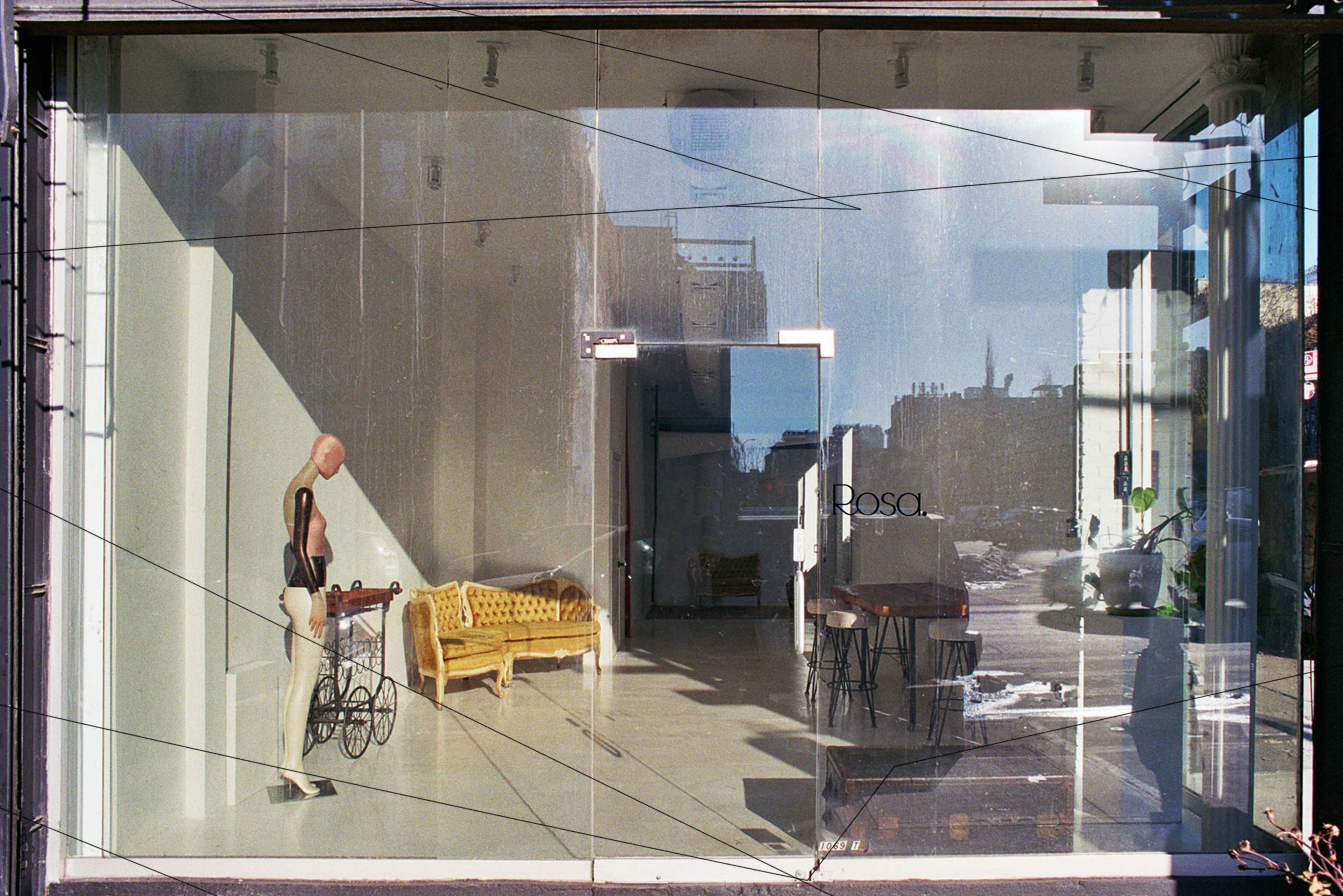 The image shows a modern storefront with large glass windows, featuring an interior space that includes a mannequin, a yellow couch, and a table, while reflections of the street and buildings outside are visible.