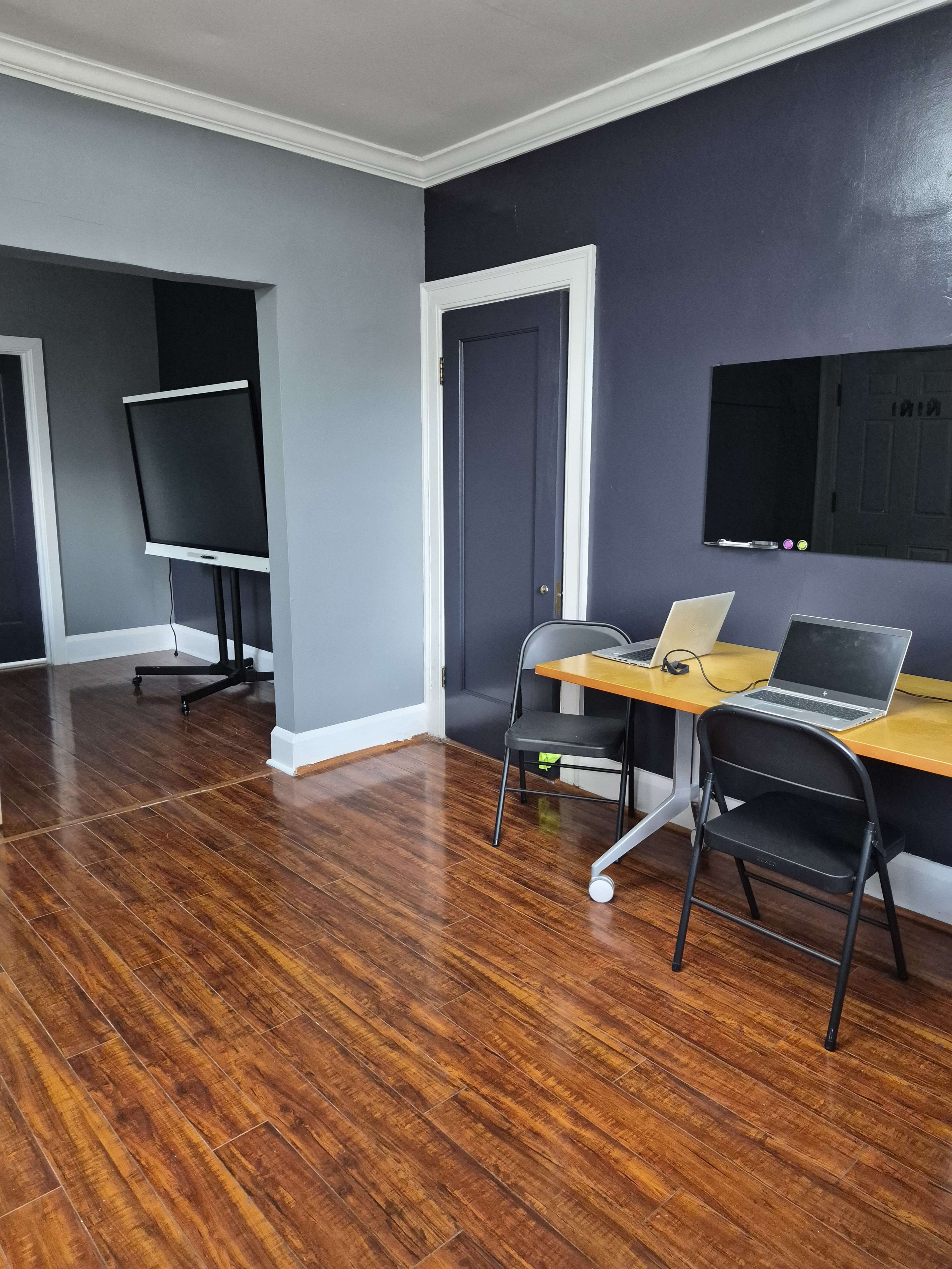The image shows a room with wooden flooring featuring a desk with two laptops, a large television on a stand, and a door leading to another space.