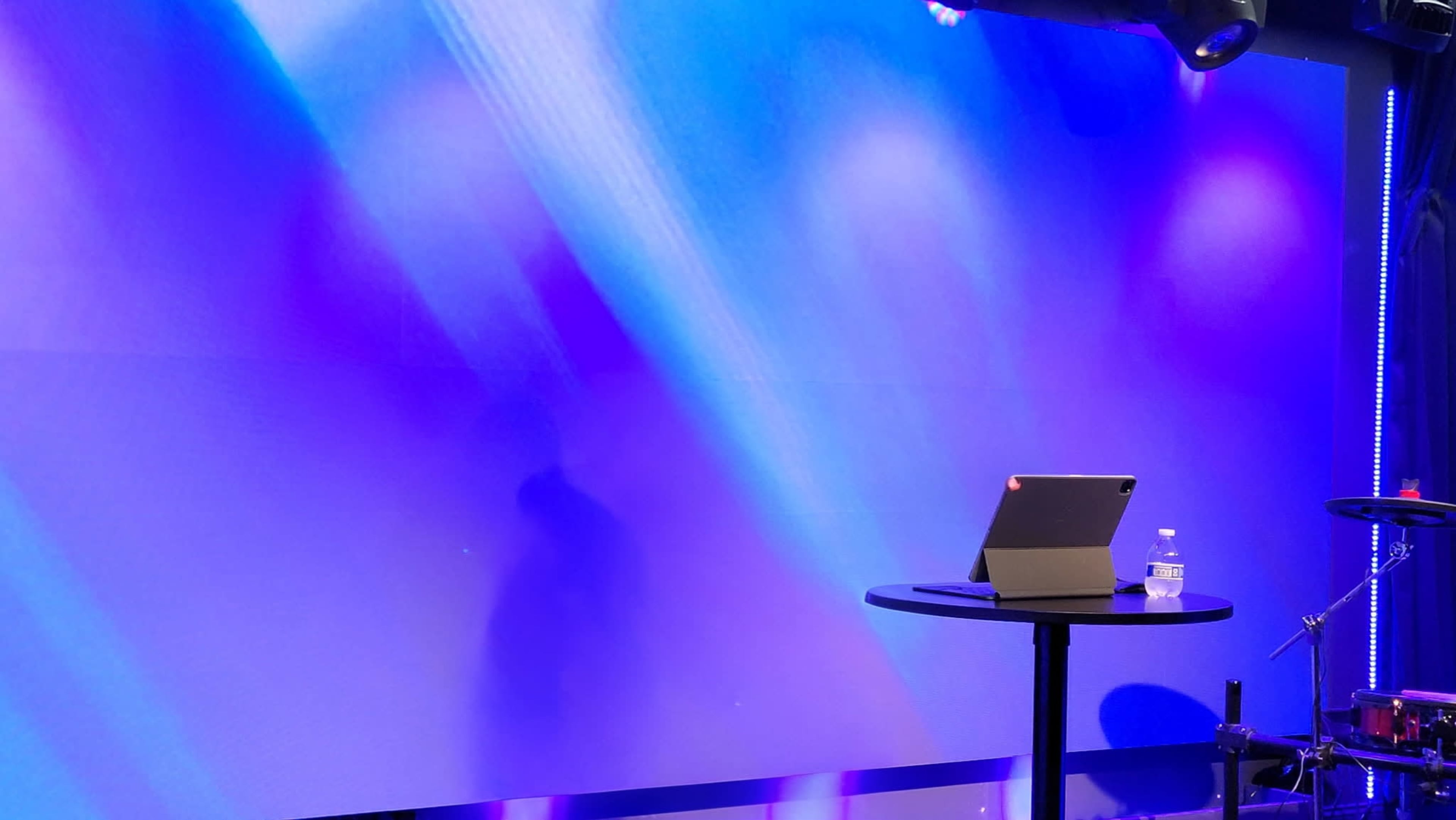 A stage is set with a purple background, featuring an iPad on a black table and a water bottle, alongside visible musical equipment.