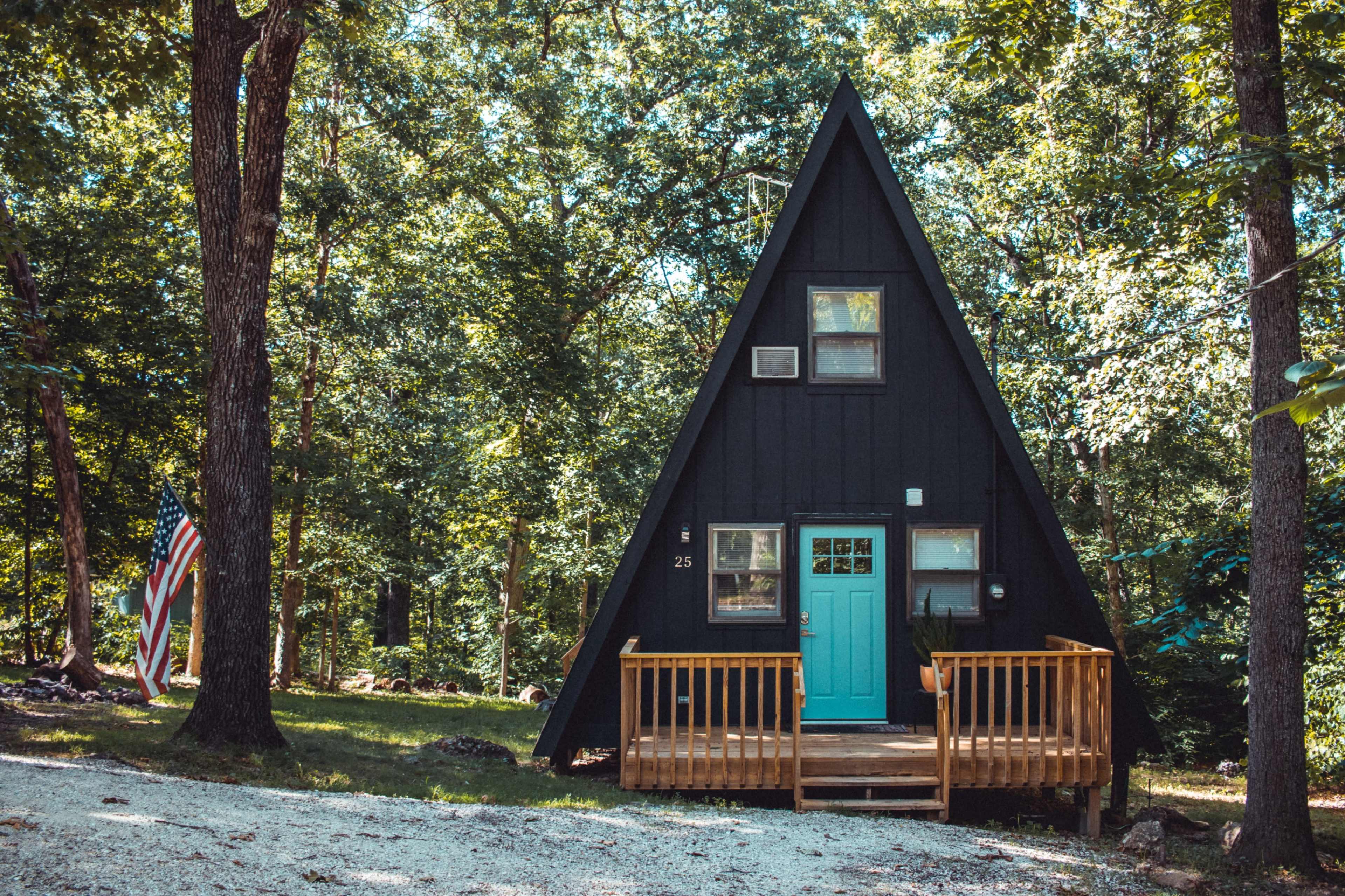 The image shows a black A-frame cabin with a turquoise door surrounded by green trees and a small wooden deck.