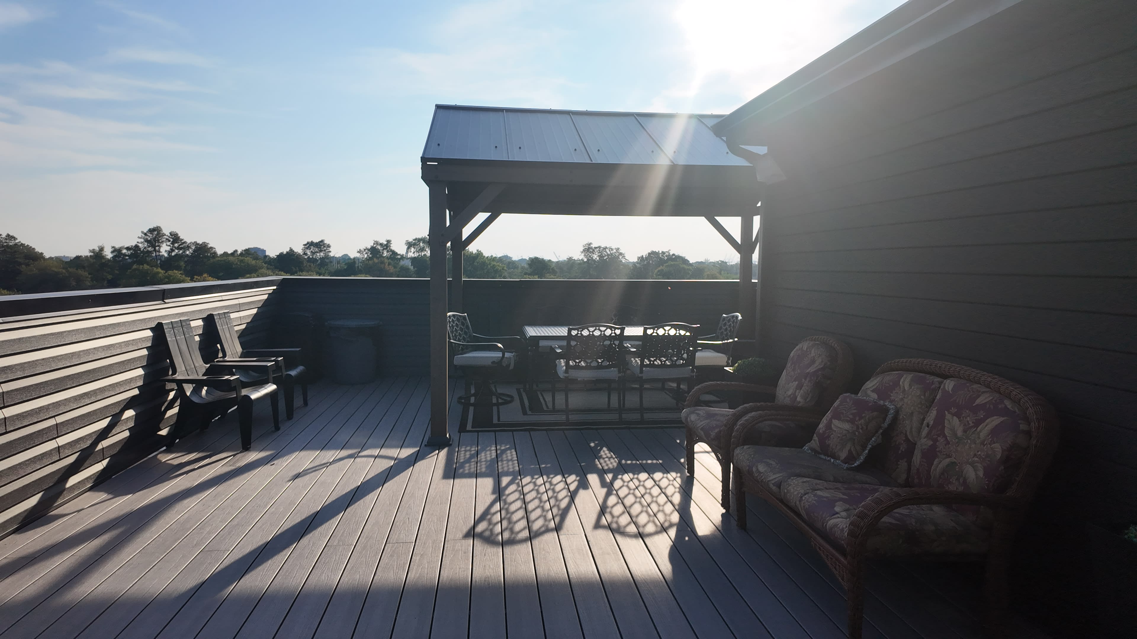 The image shows a rooftop deck with a covered seating area, including chairs and a table, under a bright sky.