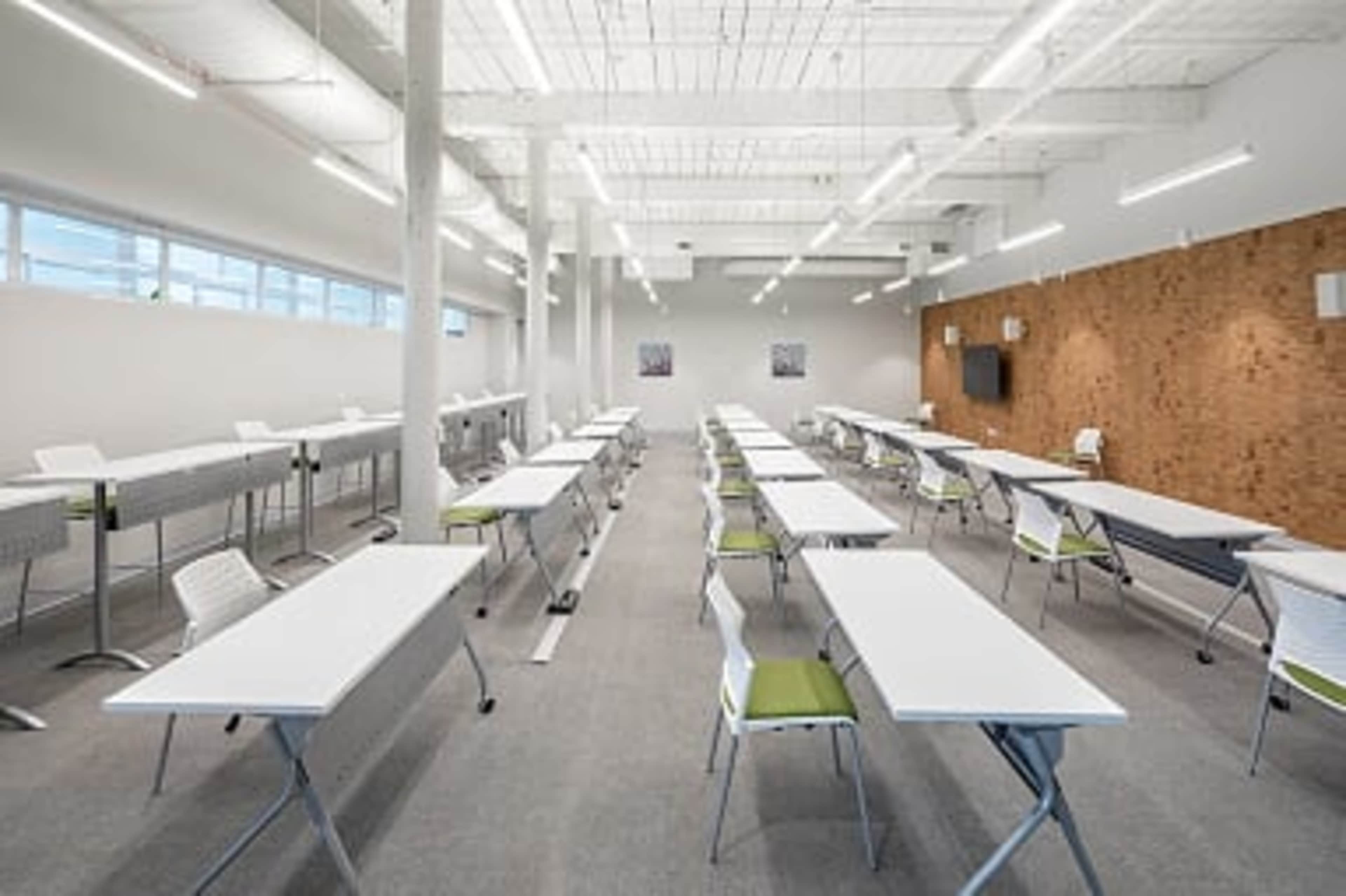 A spacious classroom arranged with long white tables and chairs on a gray carpeted floor, featuring bright overhead lighting and a brick accent wall.