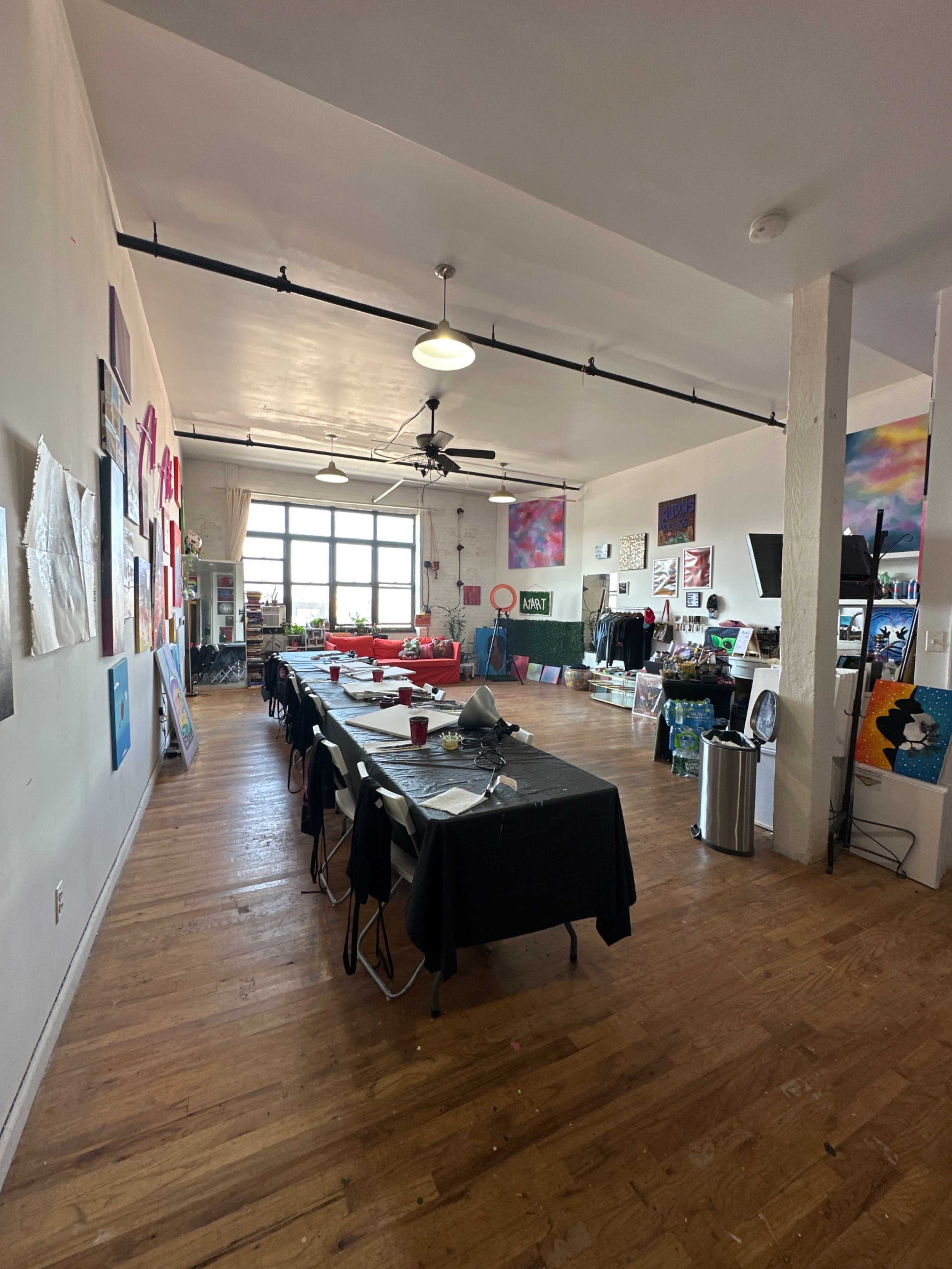 An art studio with a long table set for a workshop, surrounded by colorful artwork on the walls and large windows letting in natural light.