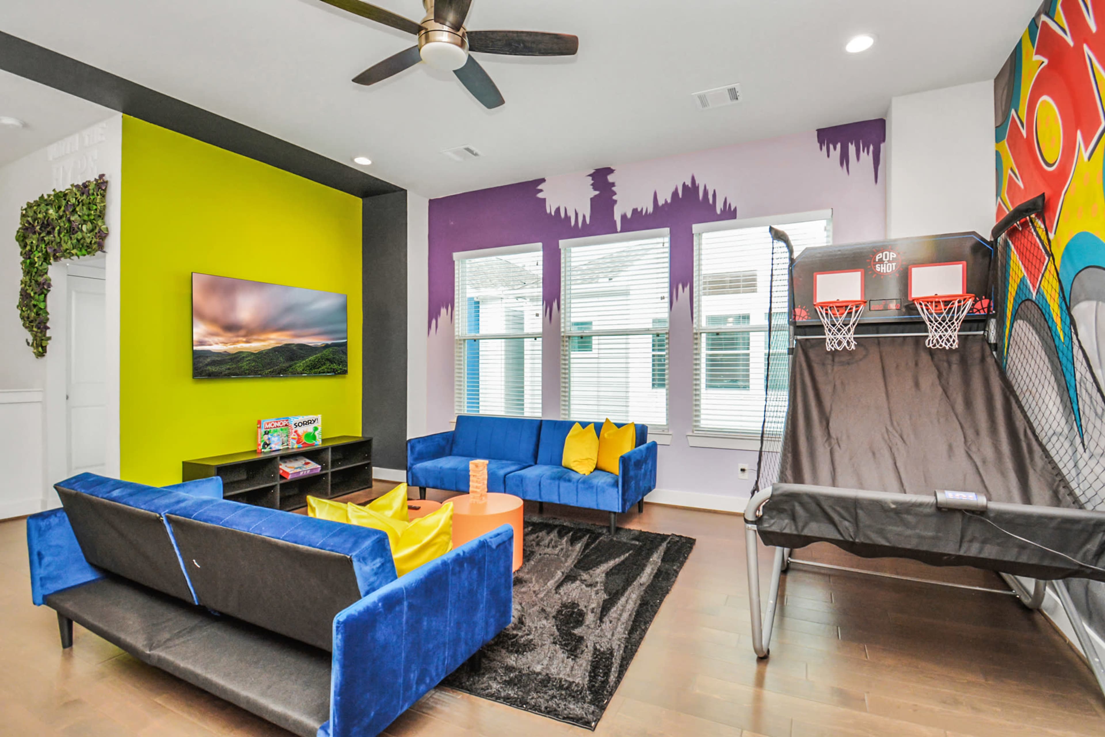 A modern living room with blue couches, a black rug, a television on a console, and a basketball shooting game against a vibrant wall.