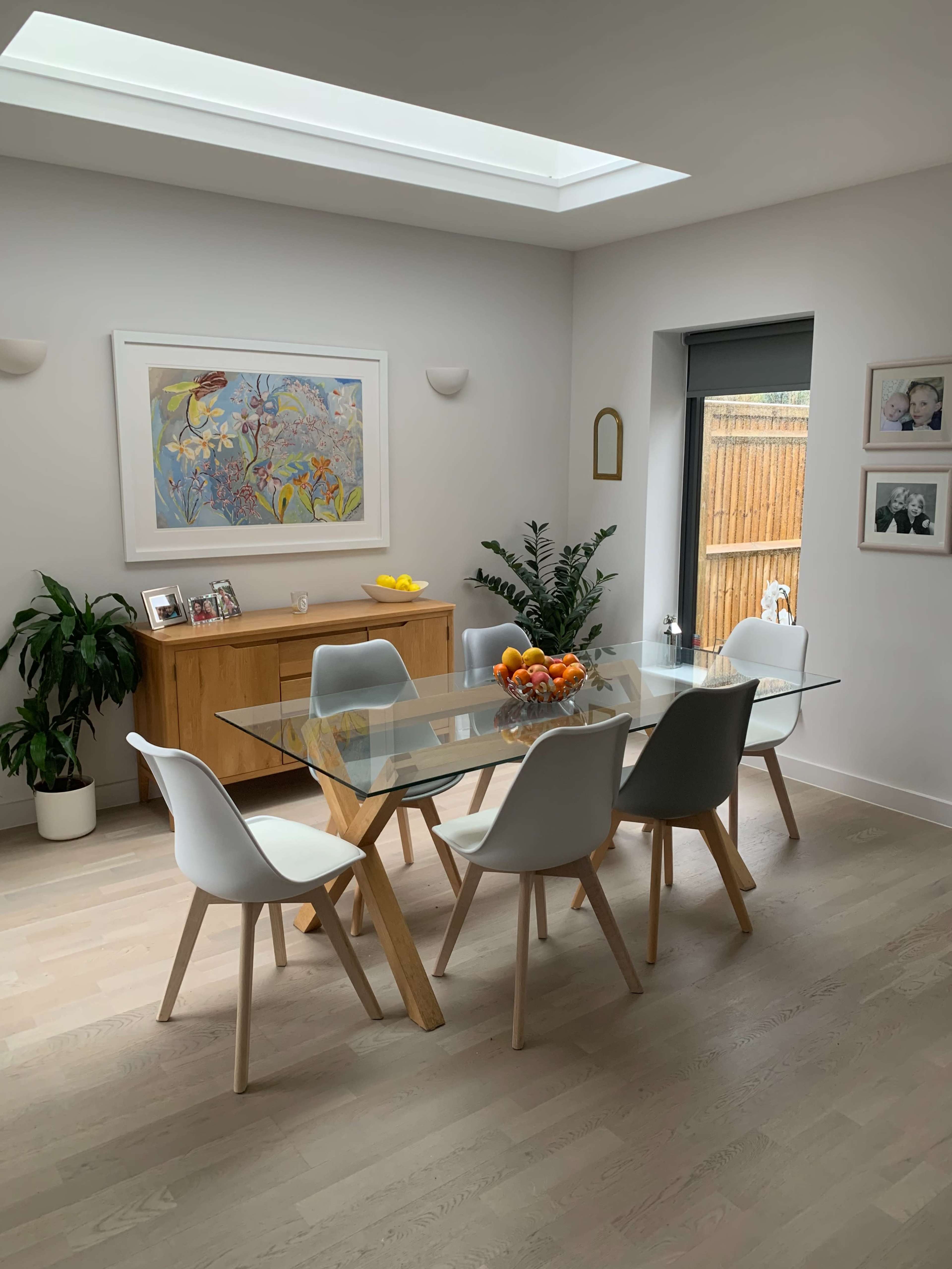 A modern dining area features a glass table surrounded by white chairs, with a wooden cabinet and plants, illuminated by natural light from a skylight.