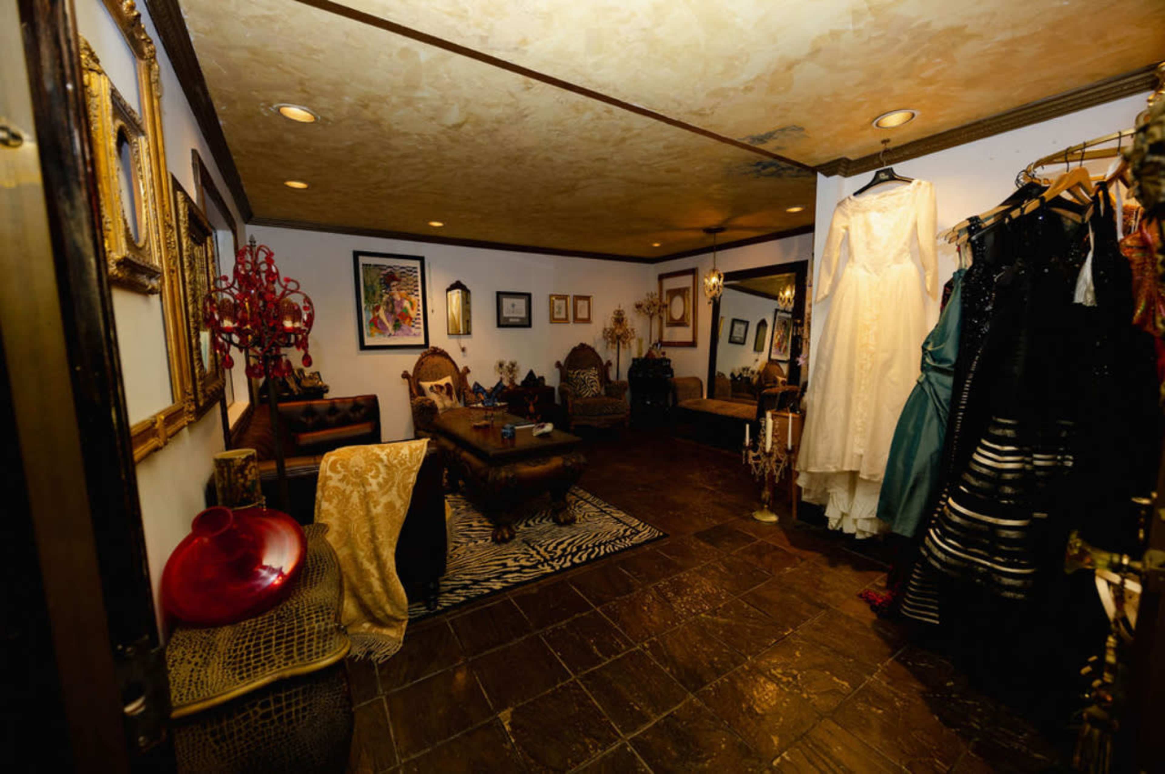 The image shows a vintage-style room adorned with ornate furniture, framed artwork, and a rack of dresses.