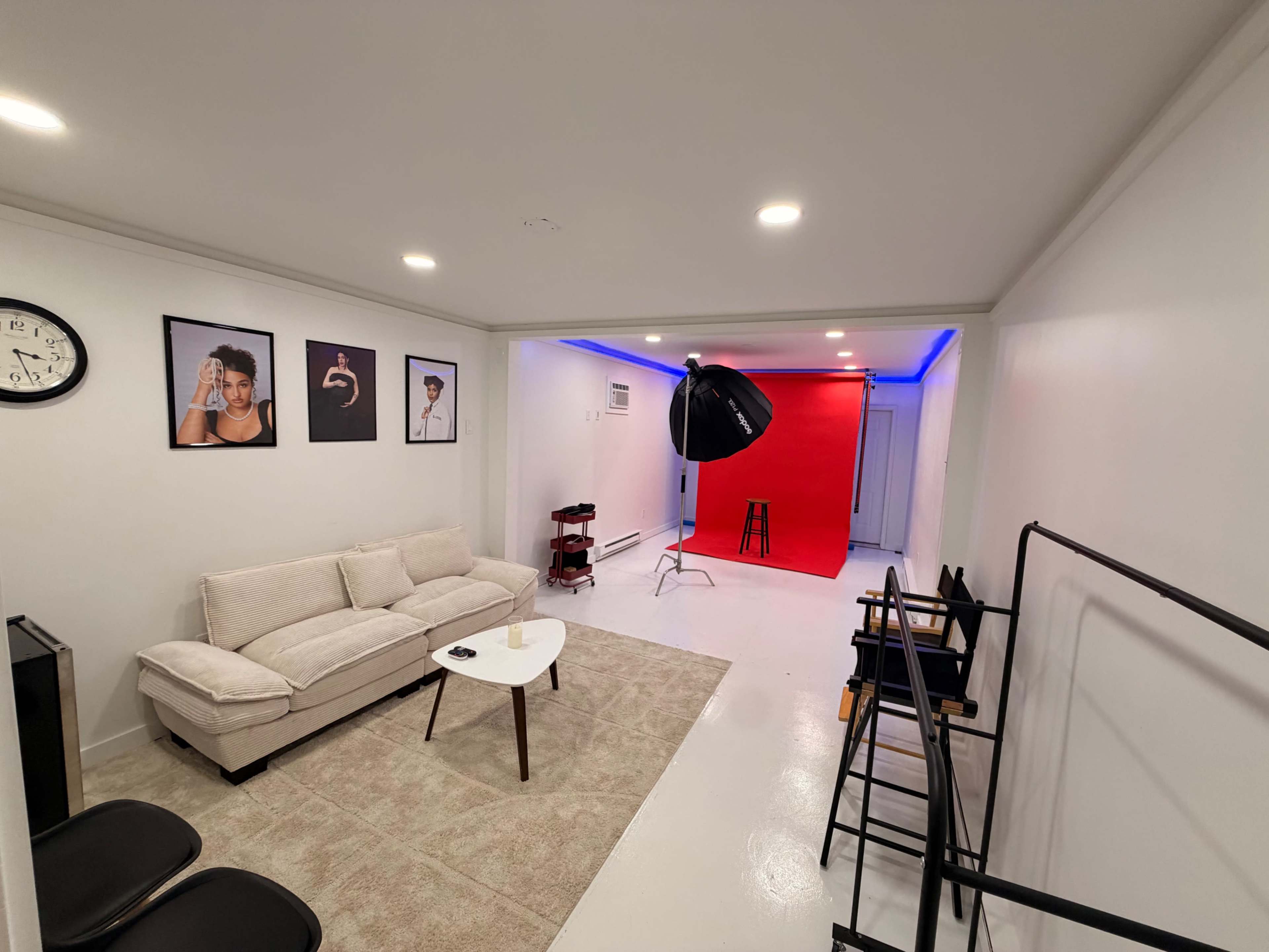 The image shows a clean, modern studio space featuring a seating area with a couch and a table, alongside a photo shoot backdrop with red lighting.