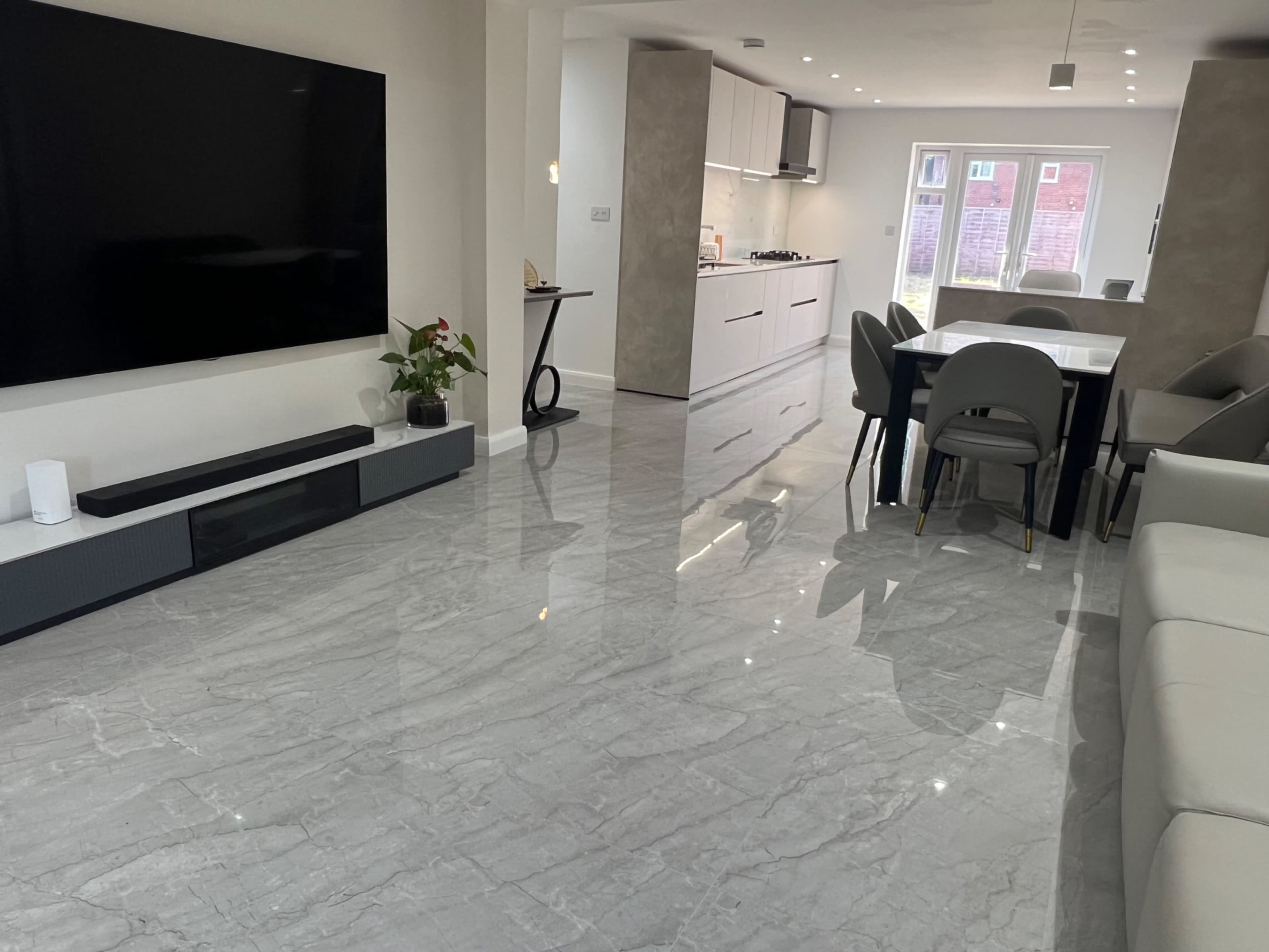 The image depicts a modern, open-concept living space featuring a large television mounted on a wall, a sleek dining table with chairs, and glossy marble flooring.