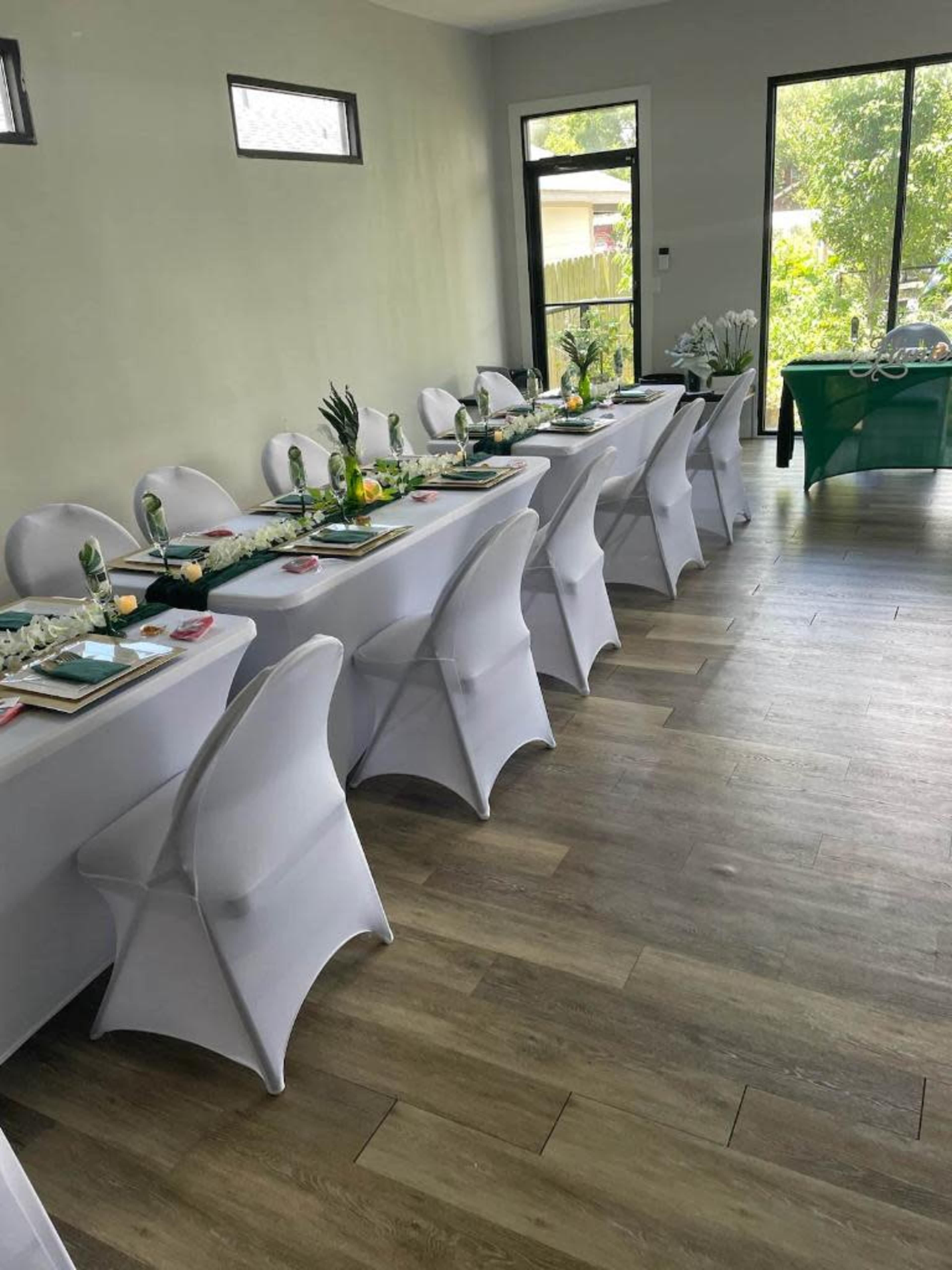 A long table is set with white tablecloths and decorations in a well-lit room, surrounded by white chairs and greenery outside the windows.