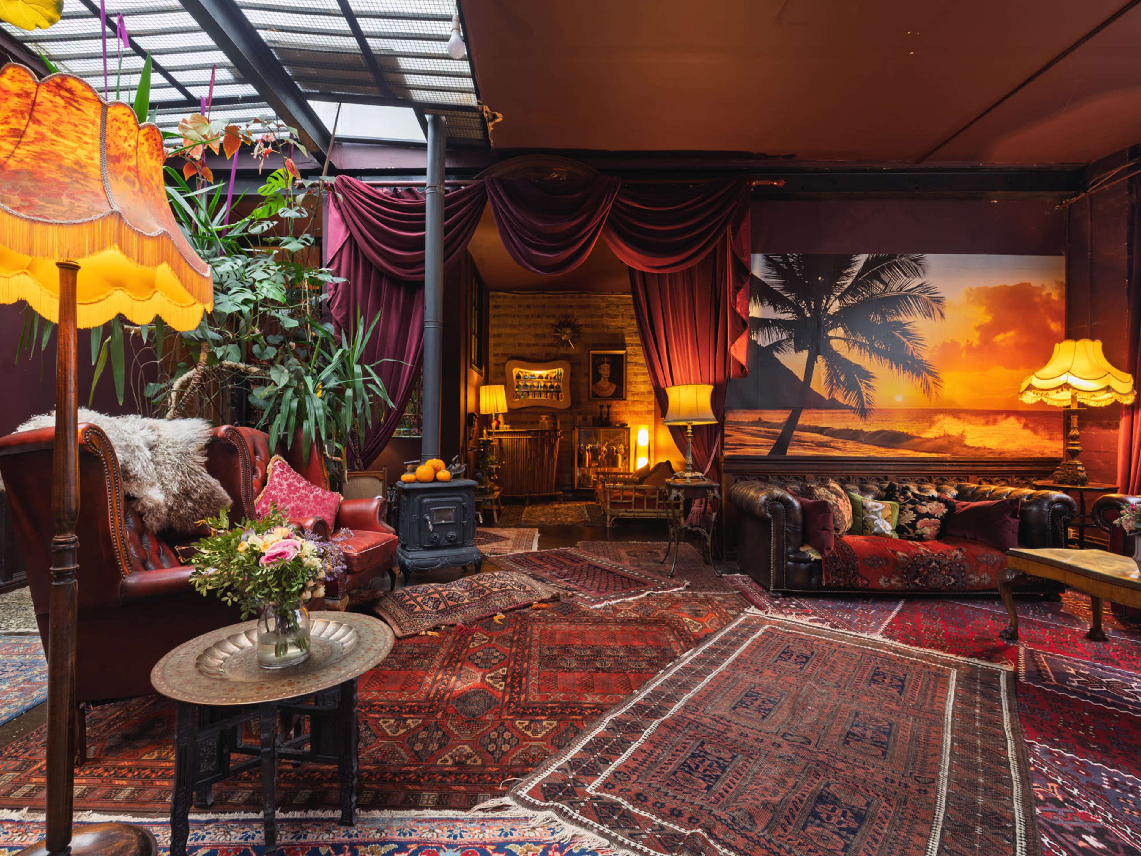 The image shows a cozy living space adorned with rich carpets, vintage furniture, and warm lighting, featuring a large wall mural of a sunset over the ocean.