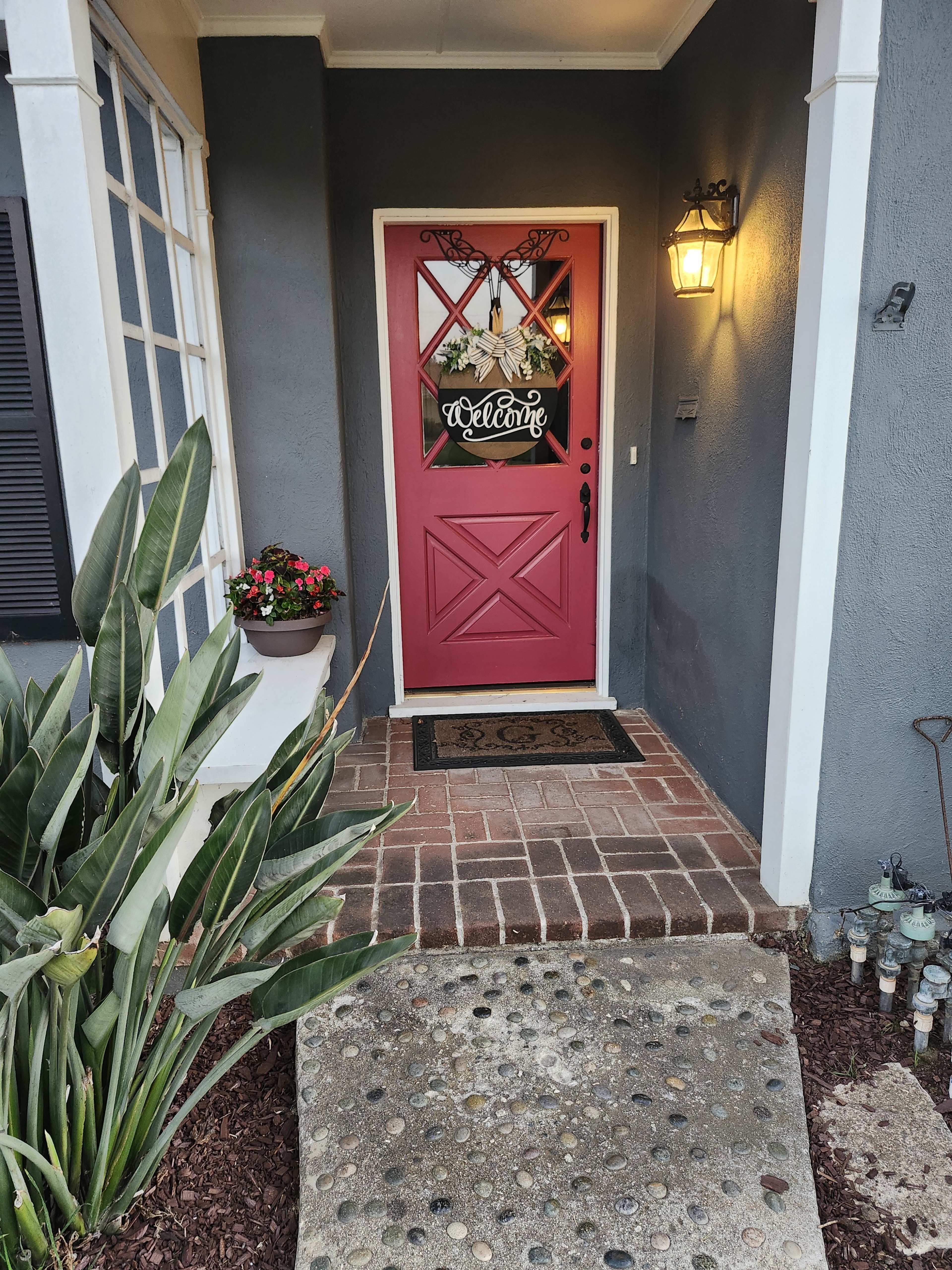 The image shows a front entryway with a pink door adorned with a welcome sign, surrounded by potted plants and a stone pathway.