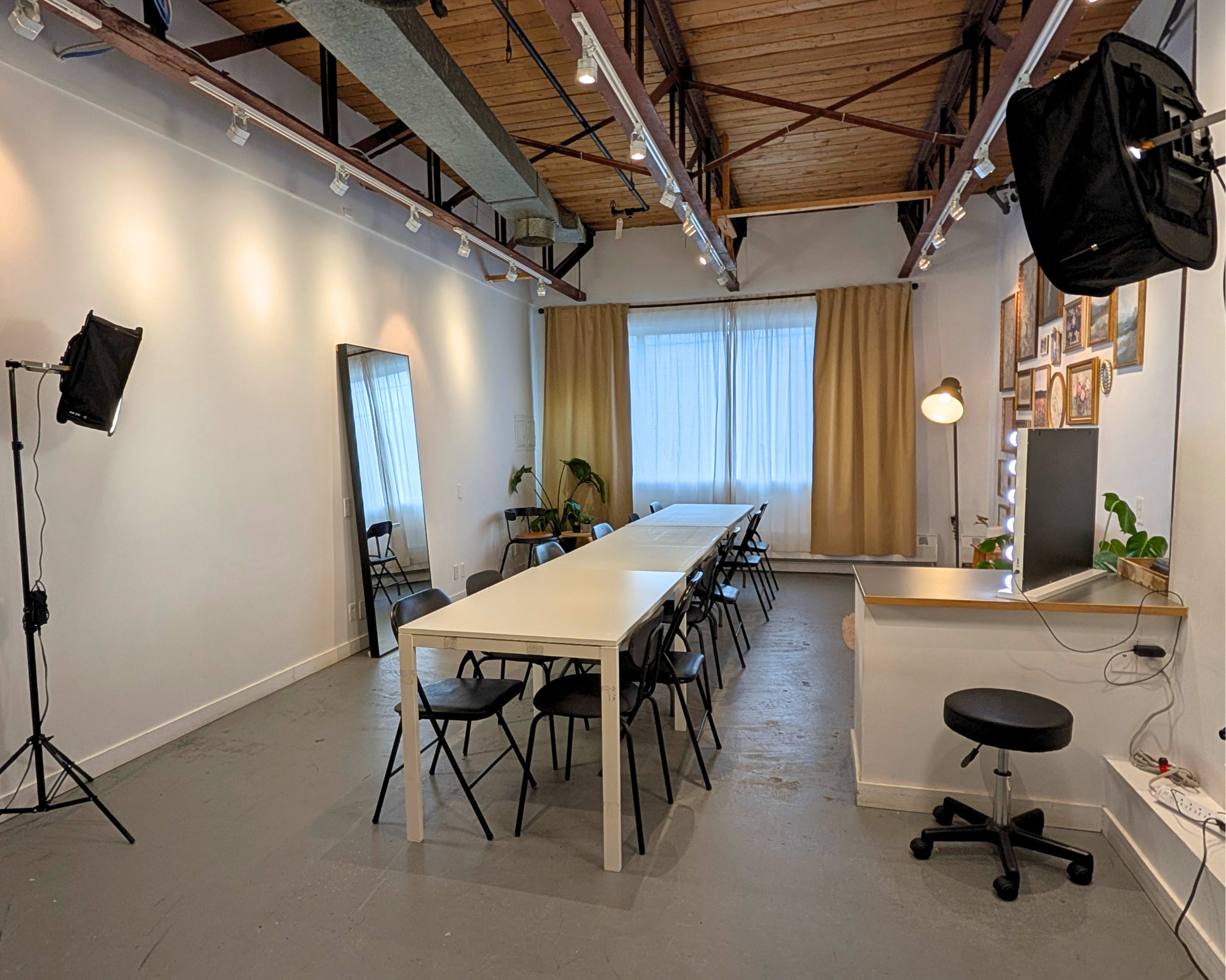 The image shows a spacious room with a long table surrounded by chairs, natural light coming through large windows, and various lighting equipment positioned around the space.