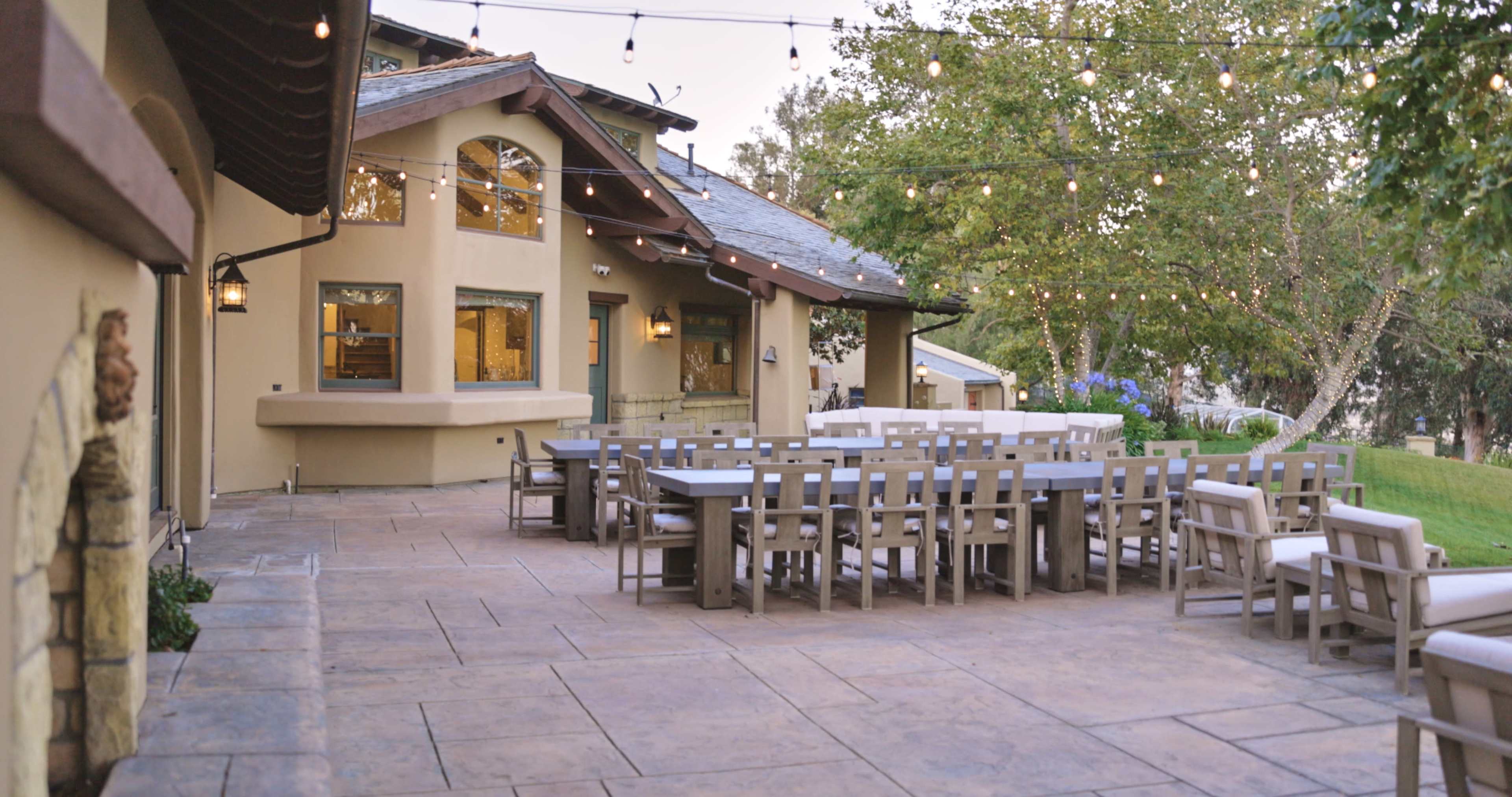 A spacious outdoor dining area with multiple tables and chairs, illuminated by string lights, adjacent to a building with large windows.
