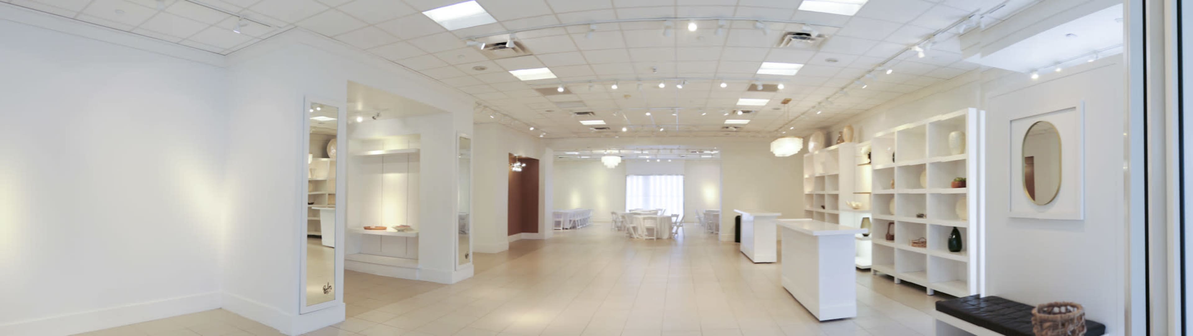 The image depicts a spacious, well-lit retail or exhibition space with white walls, display shelves, and a few tables set up for presentations or sales.