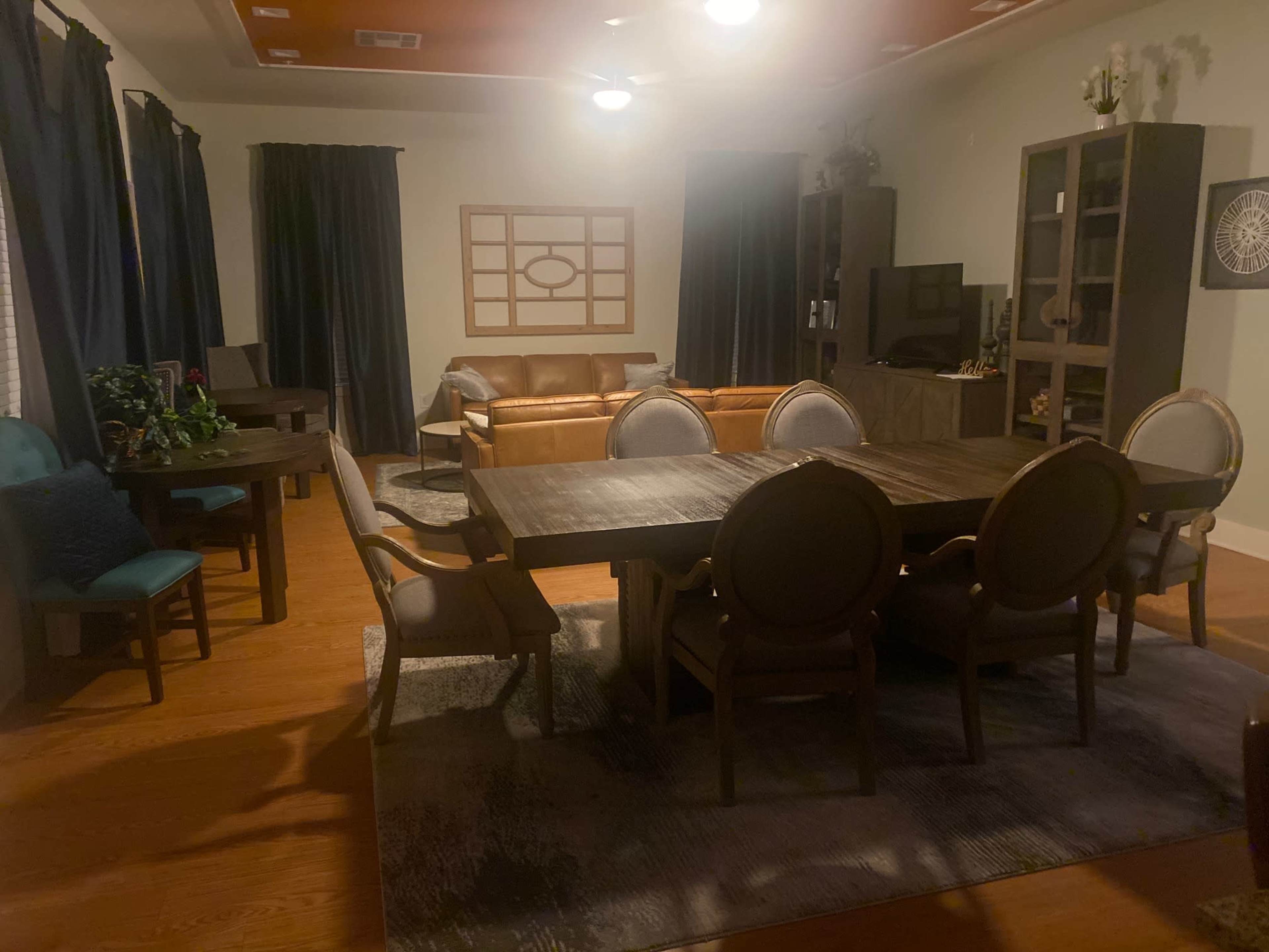 A living room features a large dining table with chairs, a seating area with a sofa, and shelves against the walls.
