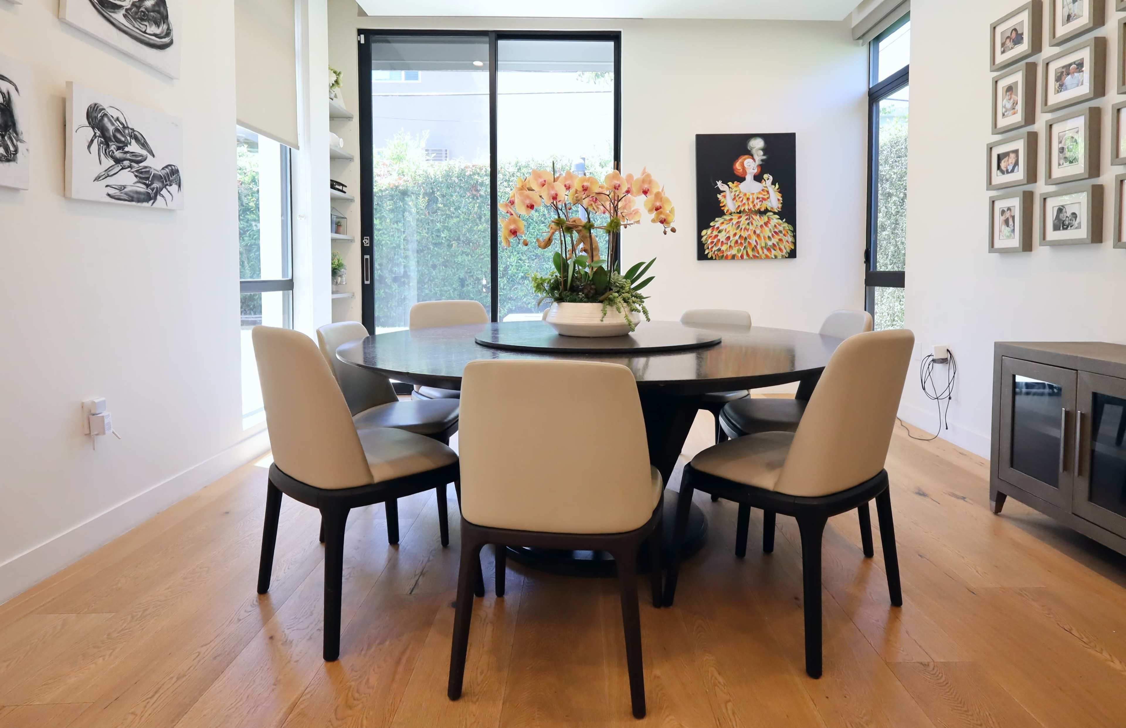 A round dining table is surrounded by six upholstered chairs in a brightly lit room featuring large windows and artwork on the walls.