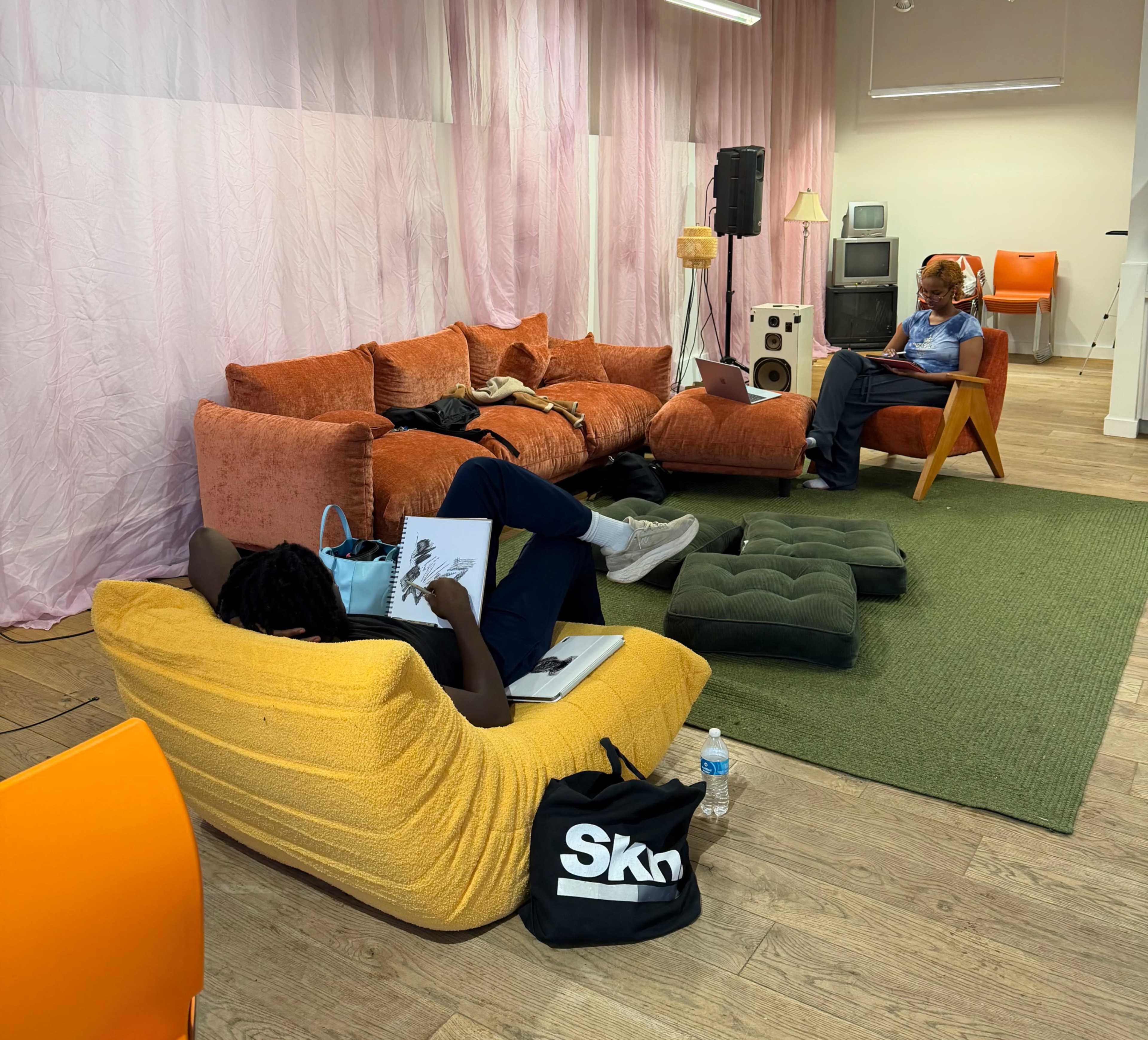 A person relaxes on a yellow bean bag while another sits on a chair, both engaging with notebooks in a spacious, modern lounge area with colorful furniture.