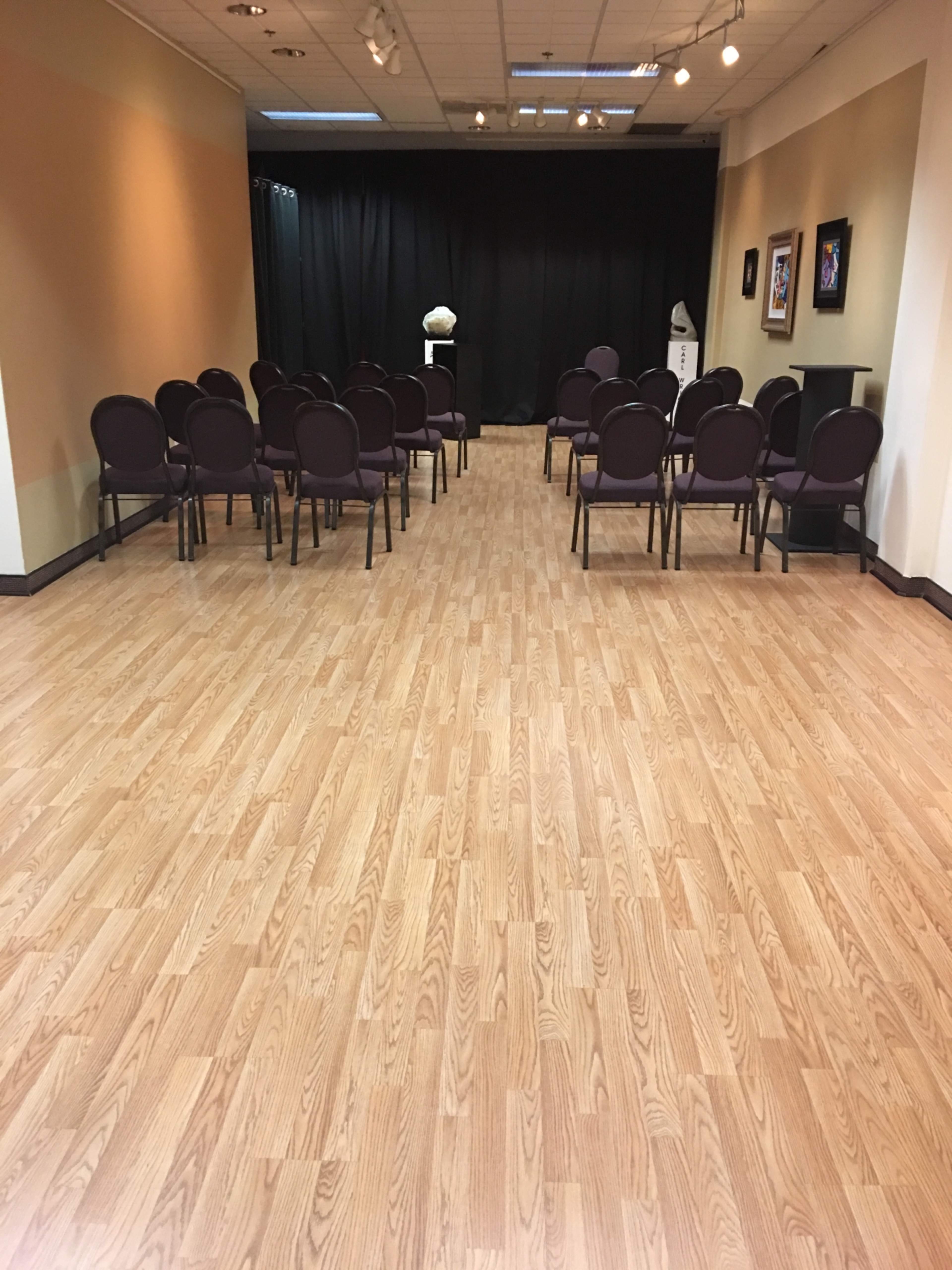A room with a polished wooden floor, arranged with several rows of purple chairs facing a black curtain and a small pedestal with a decorative item.