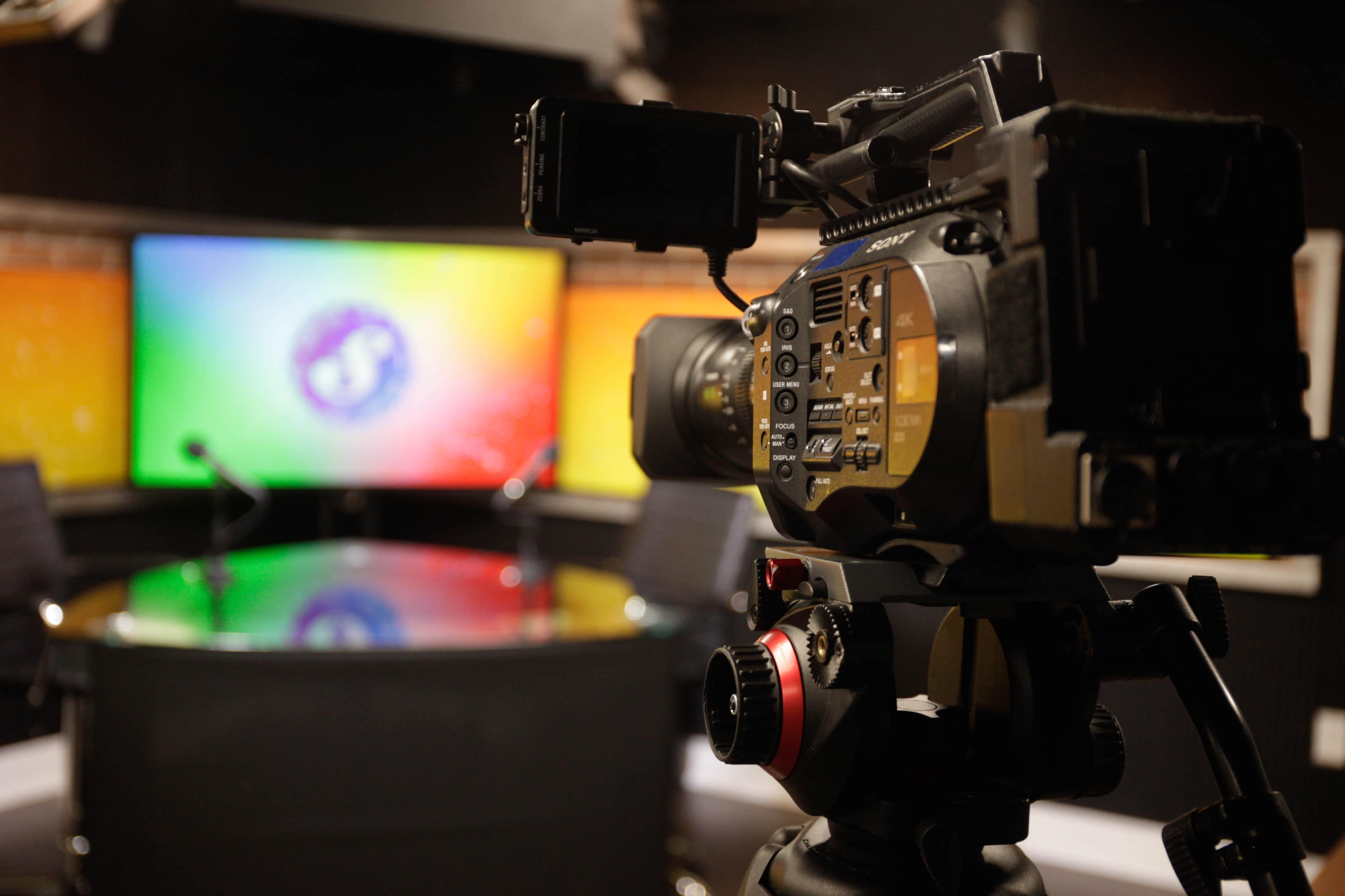 A video camera is positioned in front of a brightly lit television studio set with a circular table and a colorful backdrop.