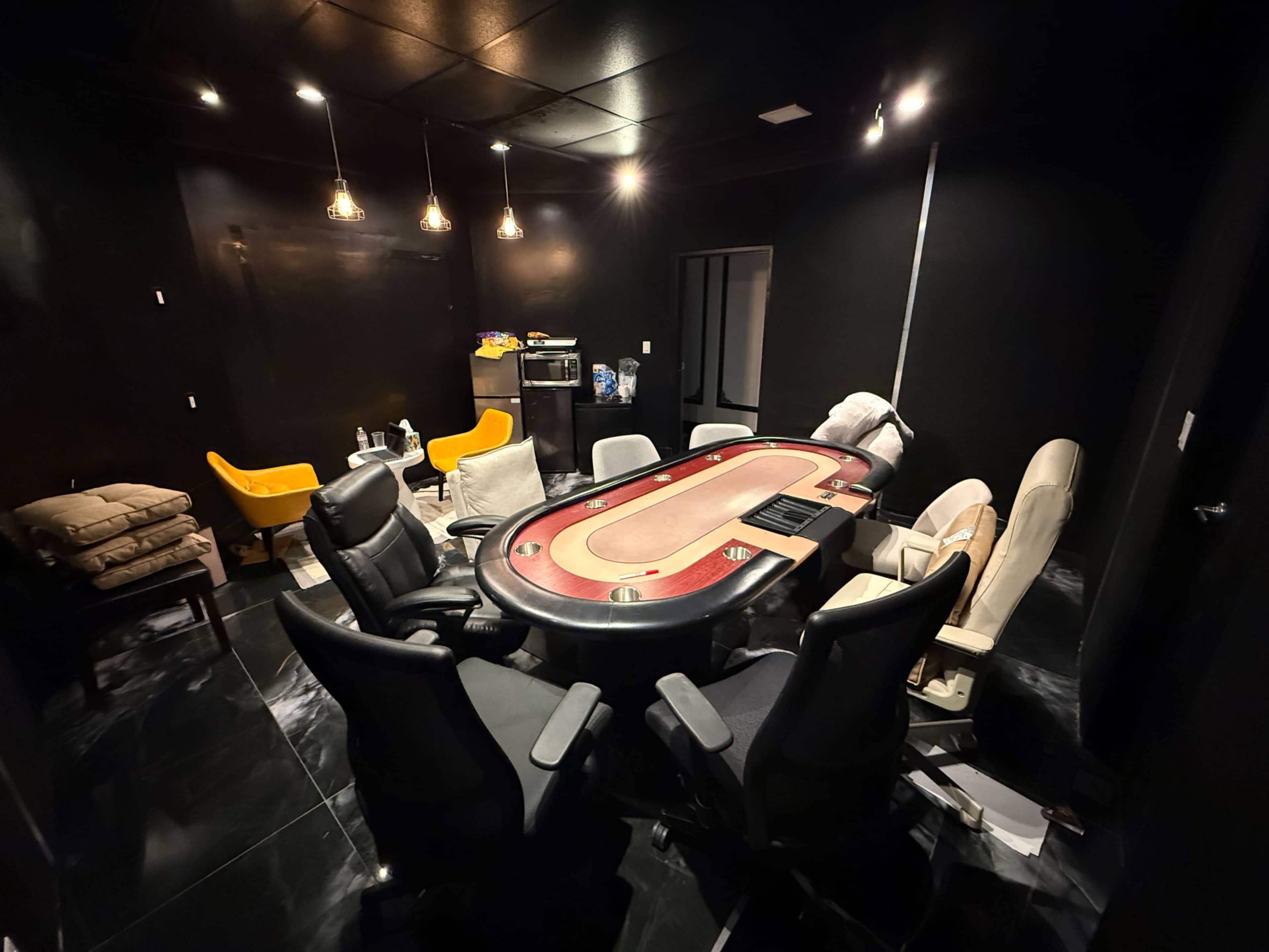 The image shows a dimly lit poker room featuring a round poker table surrounded by several chairs and yellow armchairs in the corners.