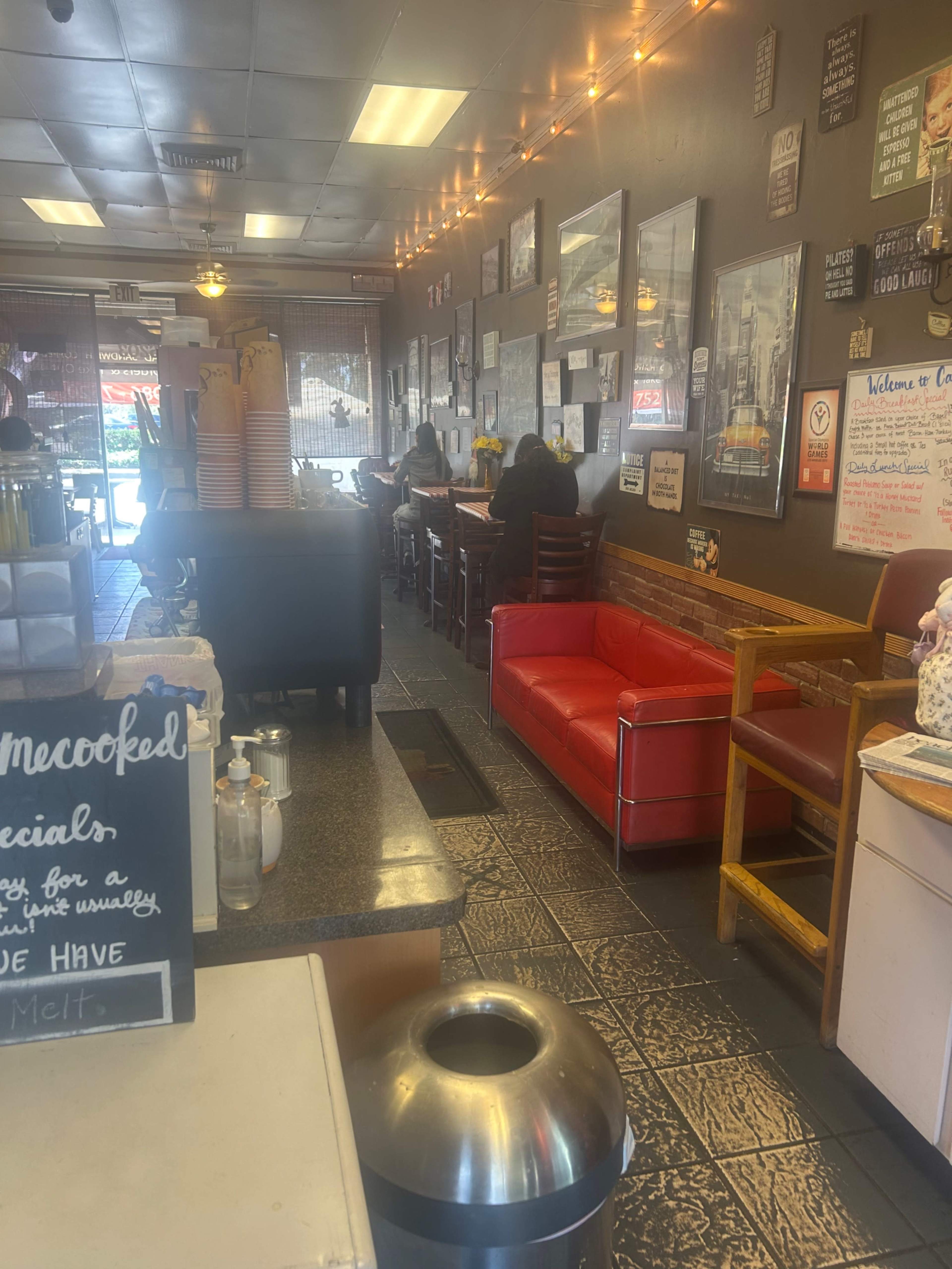 The image shows a cozy café interior with a counter for ordering, a seating area featuring red and wooden benches, and several patrons engaged in conversation.